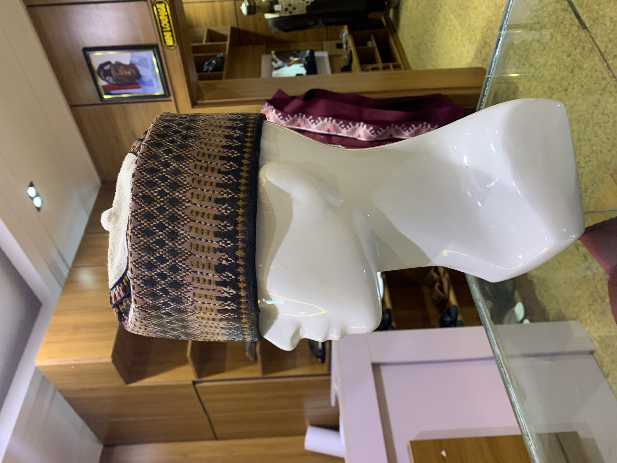 The image shows a white mannequin head wearing a woven hat with a diamond pattern and a white pom-pom. The mannequin is positioned in front of a glass display counter, and the background suggests an indoor setting that appears to be a shop or boutique. Wooden shelves and displays are visible, with some clothing items and what looks like shoes on the shelves. A framed portrait of a man in traditional attire hangs on a wooden wall in the background, along with a sign that reads "MINI LOUNGE." The lighting in the shop appears to be from overhead fixtures. The presence of clothing and display units indicates a retail environment, likely in Gura Top, Nigeria, given the context of the provided information. The overall scene depicts a retail display for traditional headwear within a Nigerian establishment.