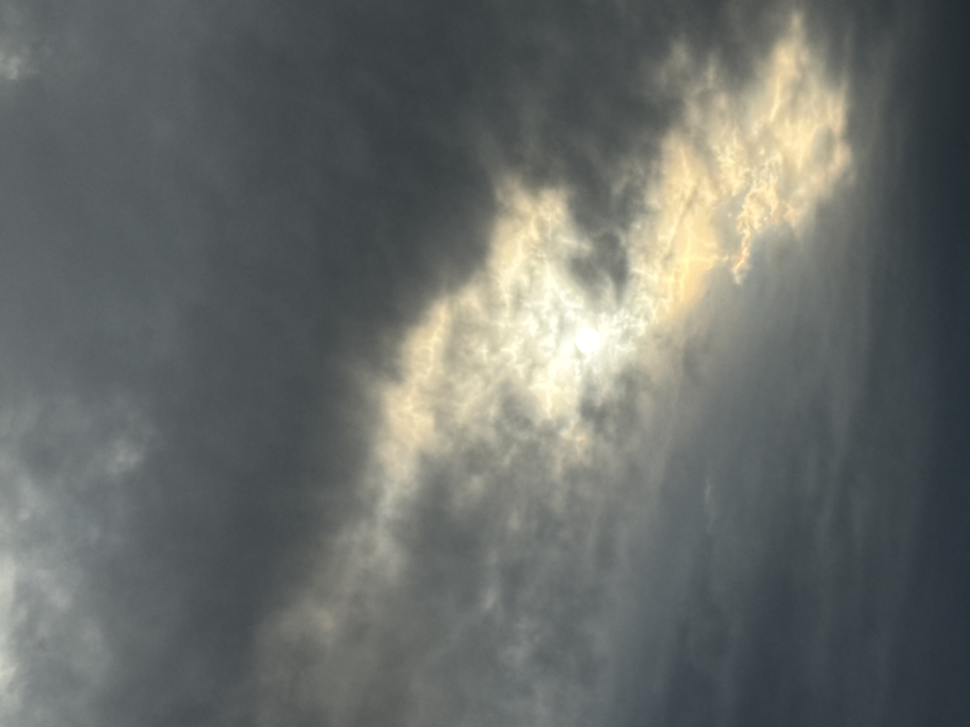 The image depicts a cloudy sky, likely during the daytime. A bright light source, possibly the sun, is partially obscured by thick, gray clouds. The light filters through a gap in the clouds, creating a contrast between the illuminated areas and the darker cloud cover. The scene suggests a weather pattern with overcast conditions and potential for a storm or heavy rain. The specific location in Chaikovsky, Russia, cannot be determined from the image alone.