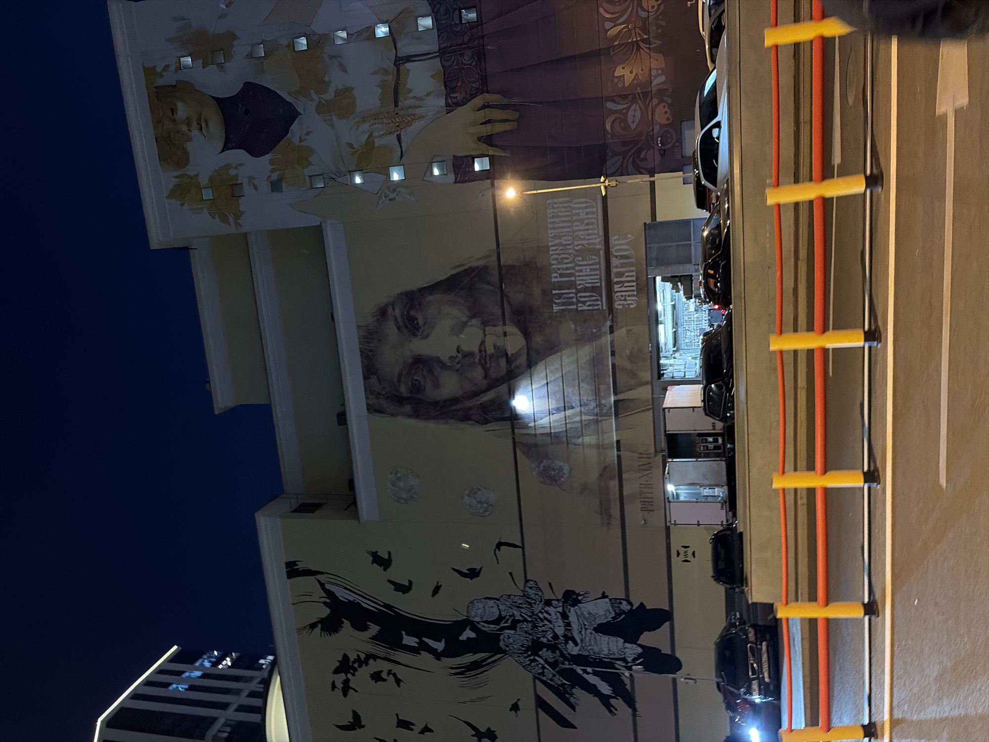 The image captures a nighttime view of a building in Moscow, Russia, adorned with large-scale murals. The building, likely a commercial or residential structure, features a modern architectural design with a prominent illuminated edge at the top. The murals are the central focus, depicting a soldier-like figure with a flock of birds on the left, and a large portrait of a woman with floral motifs on the right. The central mural displays a detailed portrait of a woman with superimposed text in Cyrillic. Below the murals, a street scene is visible with parked cars and a streetlamp illuminating part of the facade. Yellow and orange barriers line the street in the foreground. The overall atmosphere is dark, with artificial lighting from the building and street creating highlights and shadows on the artworks and surrounding environment.