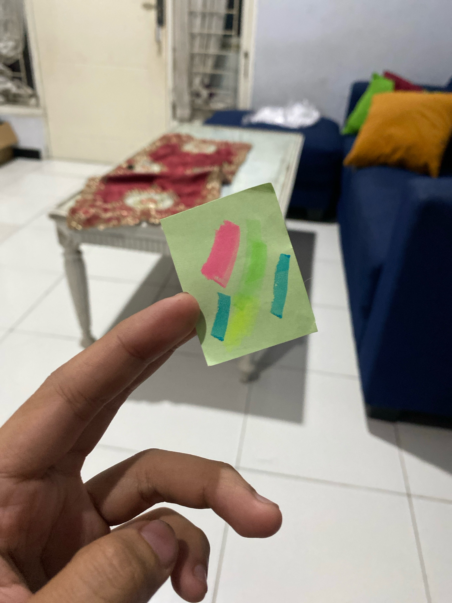 A close-up, low-angle shot shows a person's hand holding a small, light green sticky note with abstract crayon or marker strokes in pink, light green, and blue. The sticky note is positioned in the foreground, obscuring a portion of the hand. In the background, a white table with a red and gold patterned runner is visible, along with a portion of a dark blue sofa and an orange cushion. The floor appears to be tiled. The scene is indoors.