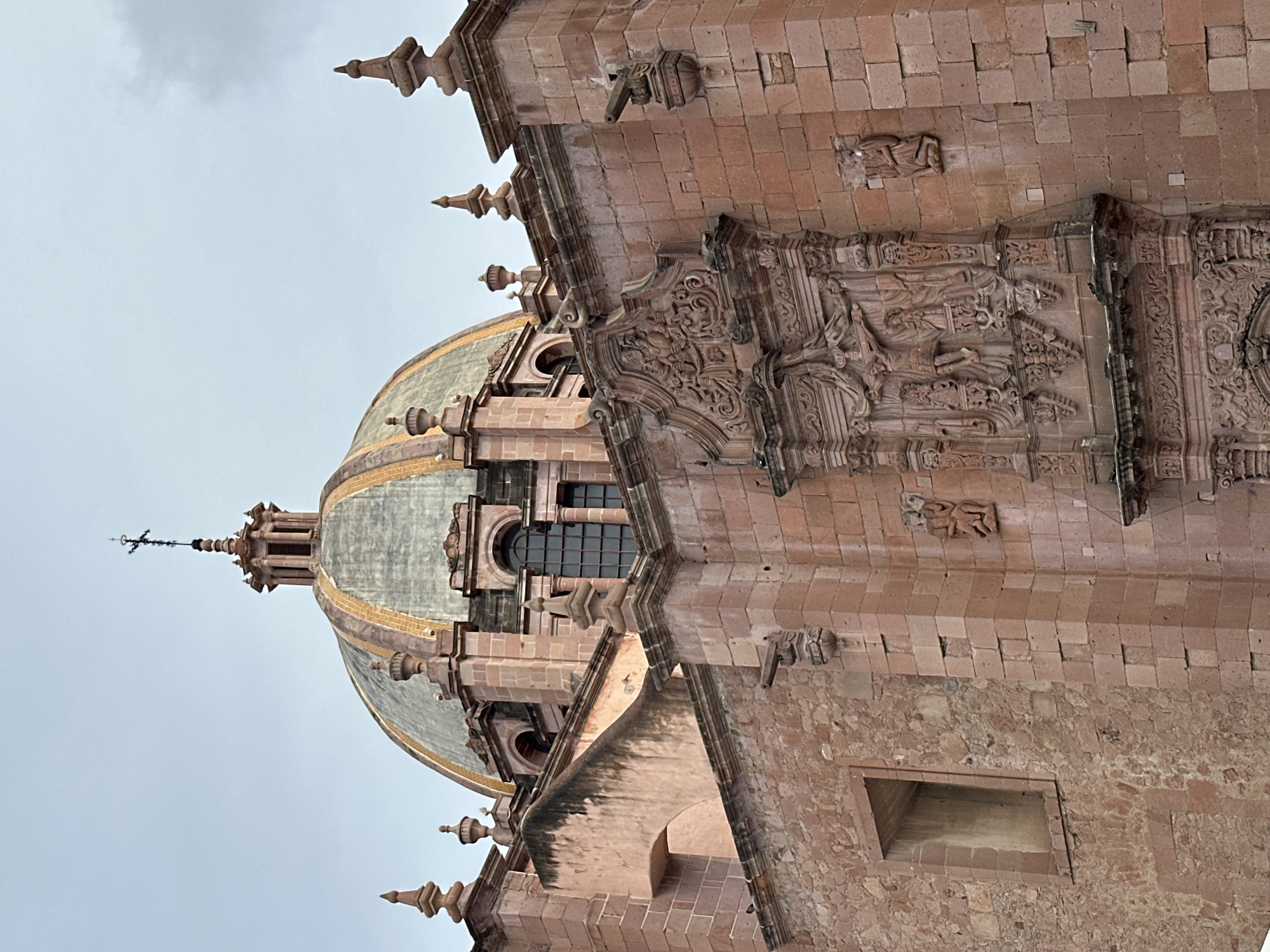 The image shows a close-up, low-angle view of the pink stone facade of the Zacatecas Cathedral in Zacatecas City, Mexico. The cathedral's prominent dome, partially obscured by the surrounding structure, is visible at the top, topped with a cross. Intricate stone carvings, including a crucifixion scene and religious figures, adorn the facade. Decorative elements like spires, gargoyles, and arched windows are also present. The sky is overcast, casting a soft light on the building.