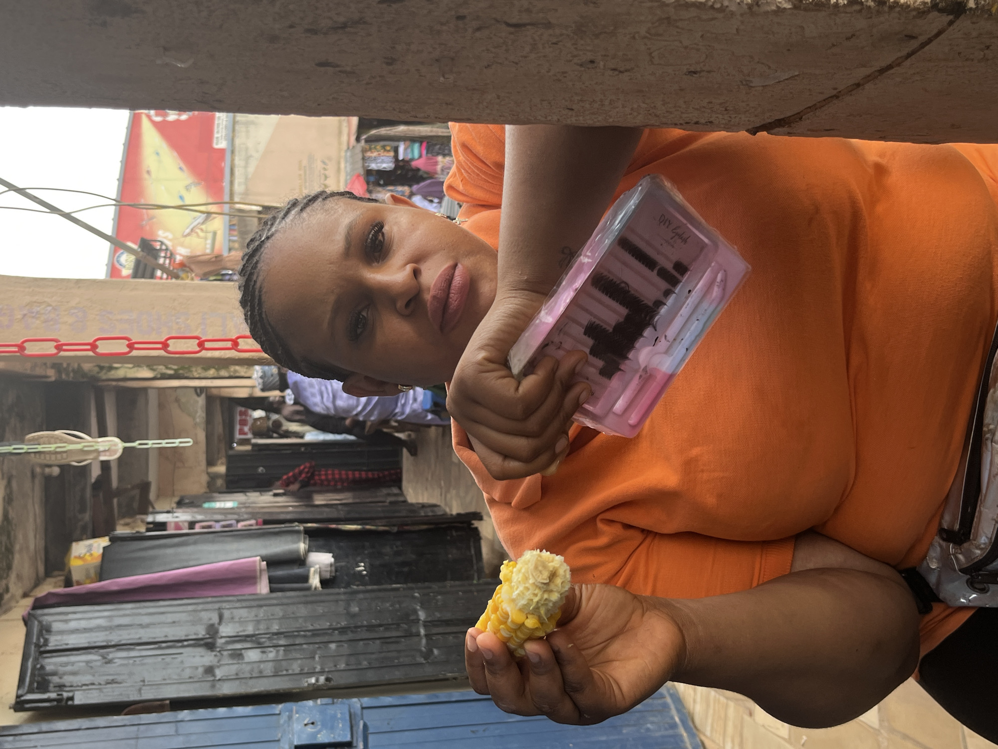 In an outdoor market scene in Jos, Nigeria, a woman with braided hair, wearing an orange t-shirt, holds a cob of corn in her left hand and a package of false eyelashes in her right. She is positioned in the foreground, with a weathered concrete pillar on the right side of the frame partially obscuring her. Behind her, a bustling market environment is visible. To the left, dark metal doors and a purple cloth are present. Further back, the market extends with more stalls, people in casual attire, and a colorful advertisement on a building facade. The lighting suggests it is daytime.