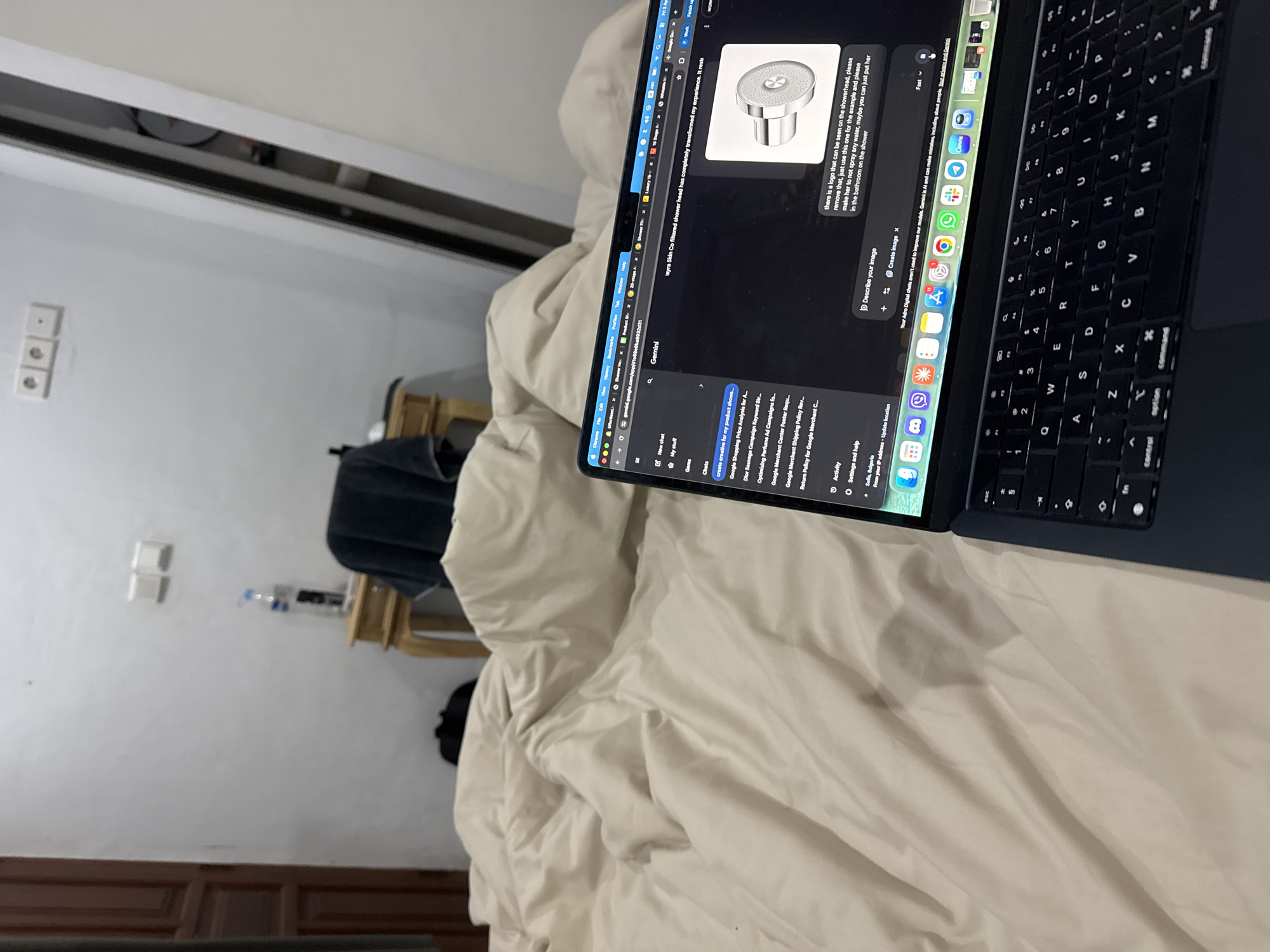 The image shows a laptop on a bed. The laptop screen displays a Google Gemini page, where the user is asking the AI to describe an image and to remove a logo from a showerhead image. The laptop keyboard is visible. The bed has beige linens. In the background, there's a wall with electrical outlets, a shelf, and a blurred object, possibly a bag or chair. The image appears to be taken indoors, likely in a bedroom. There is no indication of the location being Munggu, Indonesia.