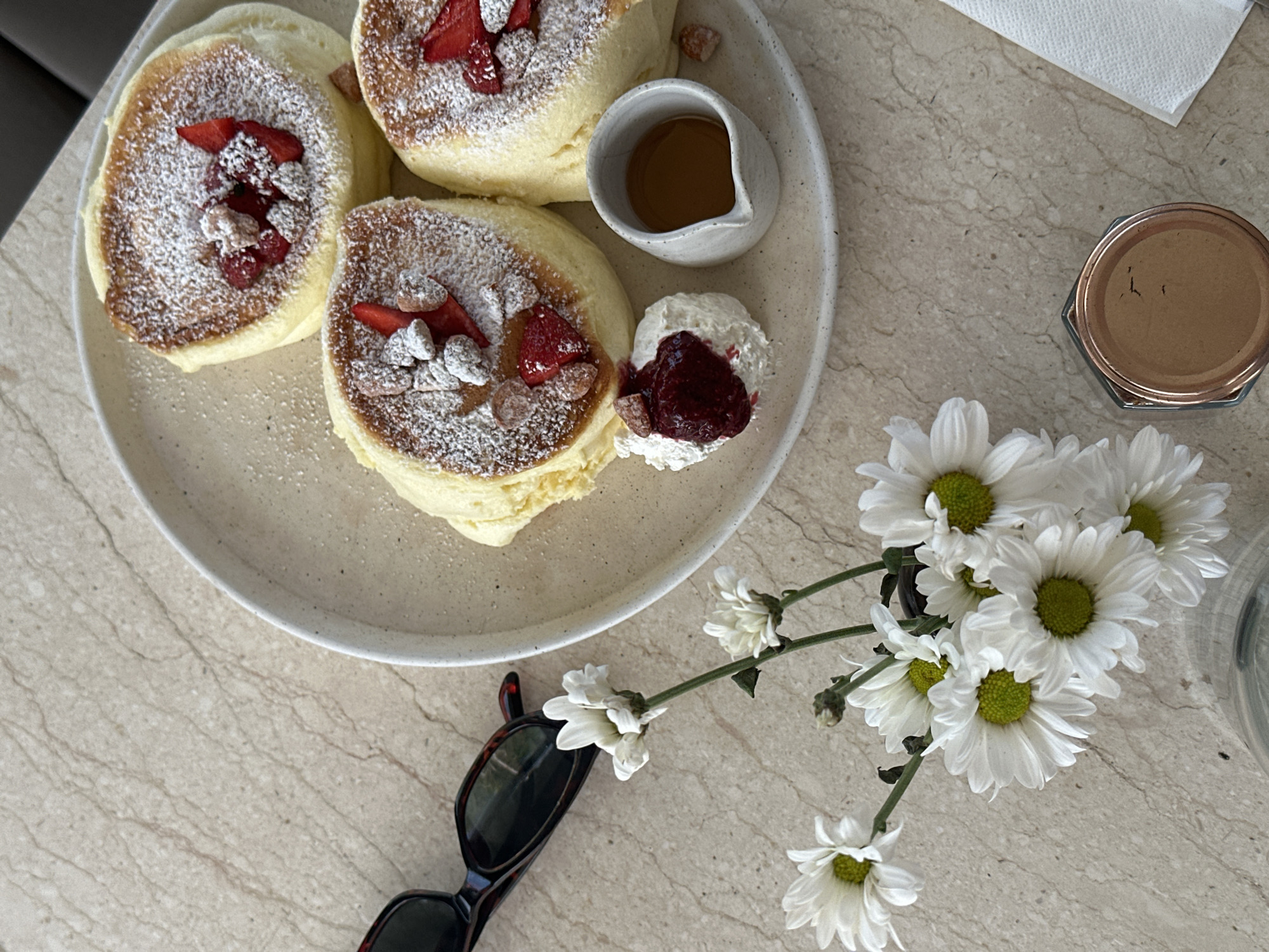 A close-up, overhead shot captures a delicate breakfast spread on a light beige, marble-patterned surface. The focal point is a light grey plate with a subtle speckled pattern, holding three fluffy, golden-yellow pancakes. Each pancake is generously dusted with powdered sugar and adorned with fresh strawberry slices and small, light brown nuts. To the side of the pancakes, a small dollop of white whipped cream is topped with a dark red berry compote and a few more nuts. Next to the cream, a small, white ceramic pitcher contains a golden-amber liquid, likely syrup or honey.
To the left of the plate, a pair of dark-rimmed sunglasses with tortoiseshell patterns rests on the surface. In the foreground and to the left, a small bouquet of white daisies with green centers adds a touch of natural beauty. A folded white paper napkin is visible in the lower right corner, and a copper-lidded glass jar is positioned below it. The background is out of focus, suggesting an indoor setting, possibly a cafe or restaurant in Mengwi, Indonesia, given the context of the query. There are no people visible in the image.