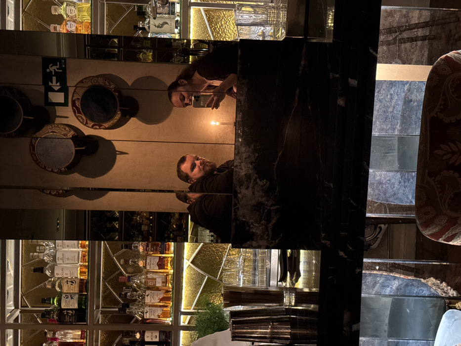 A mirror captures a bar scene with shelves filled with liquor bottles. Two people are reflected, one holding a phone, seemingly taking a picture. Wall-mounted, decorative circular objects are visible. The foreground features a dark, possibly marble, countertop and a patterned chair. The image is fragmented due to the nature of the mirrors, creating a distorted yet visually interesting composition.