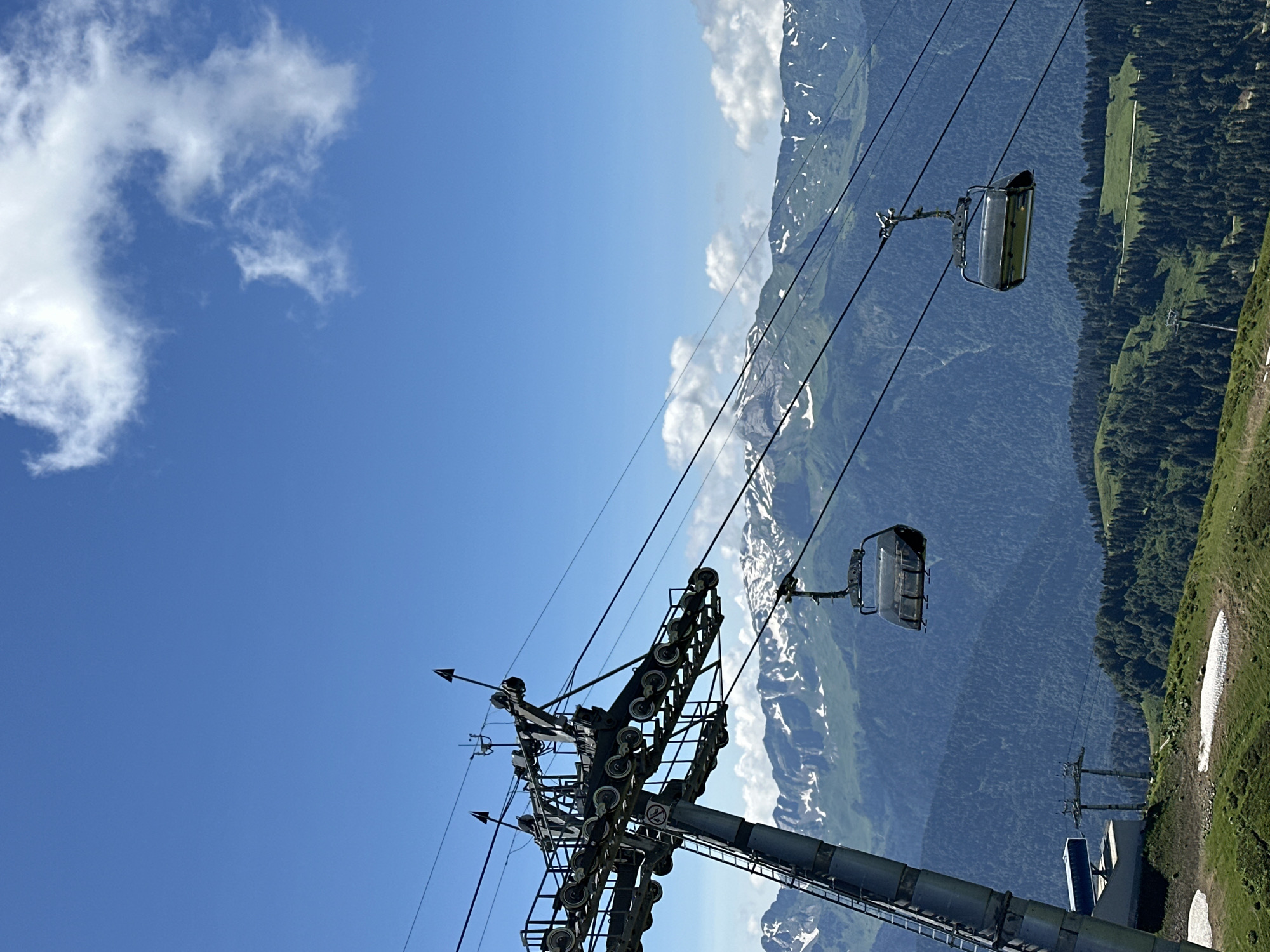 The image captures a sunny day in the resort city of Estosadok, Russia, characterized by a clear blue sky dotted with fluffy white clouds. In the foreground, a large ski lift structure dominates the left side of the frame, its metallic framework extending upwards with numerous wheels and cables. Two empty ski lift chairs are suspended from the cables, with one closer to the viewer and another further away. These chairs appear to be made of dark metal and glass.
The background reveals a majestic mountain range, with snow-capped peaks visible against the blue sky. The mountains are covered in green vegetation and dense coniferous forests on their lower slopes. A winding path or road can be seen snaking through the greenery in the mid-ground. Patches of snow are visible on the grassy slopes in the lower part of the image, suggesting it is either early summer or late spring. The overall impression is one of a picturesque mountain landscape, likely a popular tourist destination for outdoor activities. There are no people visible in the image.