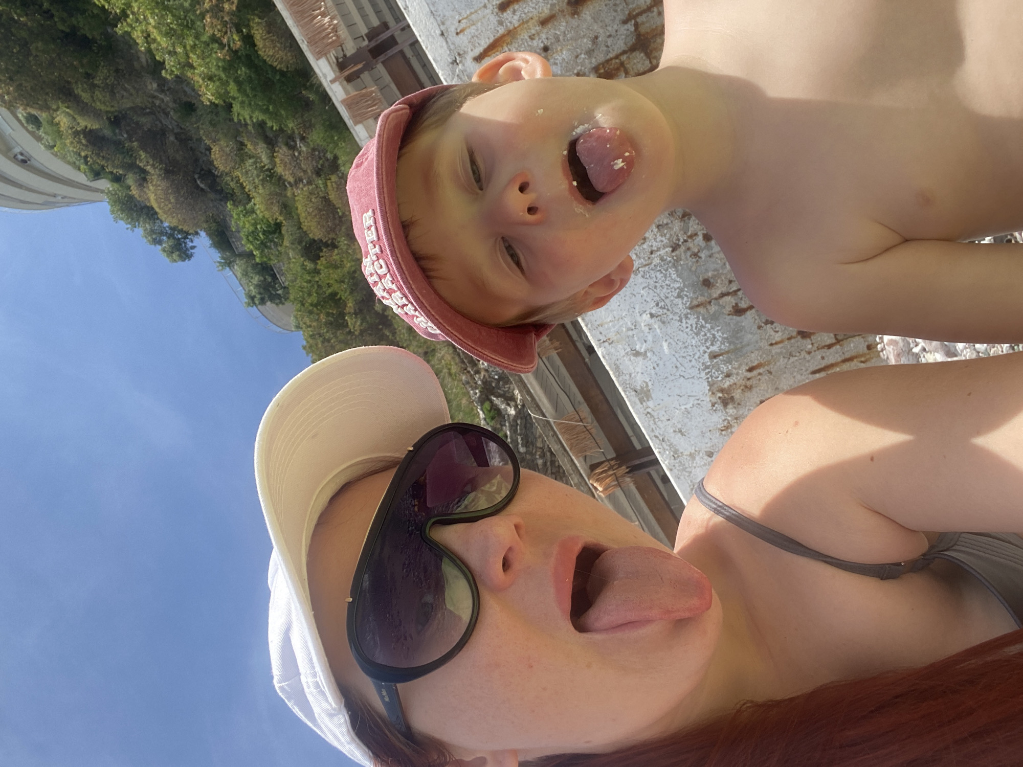 A photo taken in Boreti, Montenegro depicts a woman and child on a beach. The woman is at the bottom of the frame, wearing a white hat and large sunglasses, with red hair visible. She has her tongue out. The child is above her, wearing a pink cap, also sticking their tongue out with some food residue visible around their mouth. Both are shirtless. The background shows trees and a building on a hillside, with a clear blue sky visible in the upper left. The foreground is a textured surface, possibly concrete or stone.