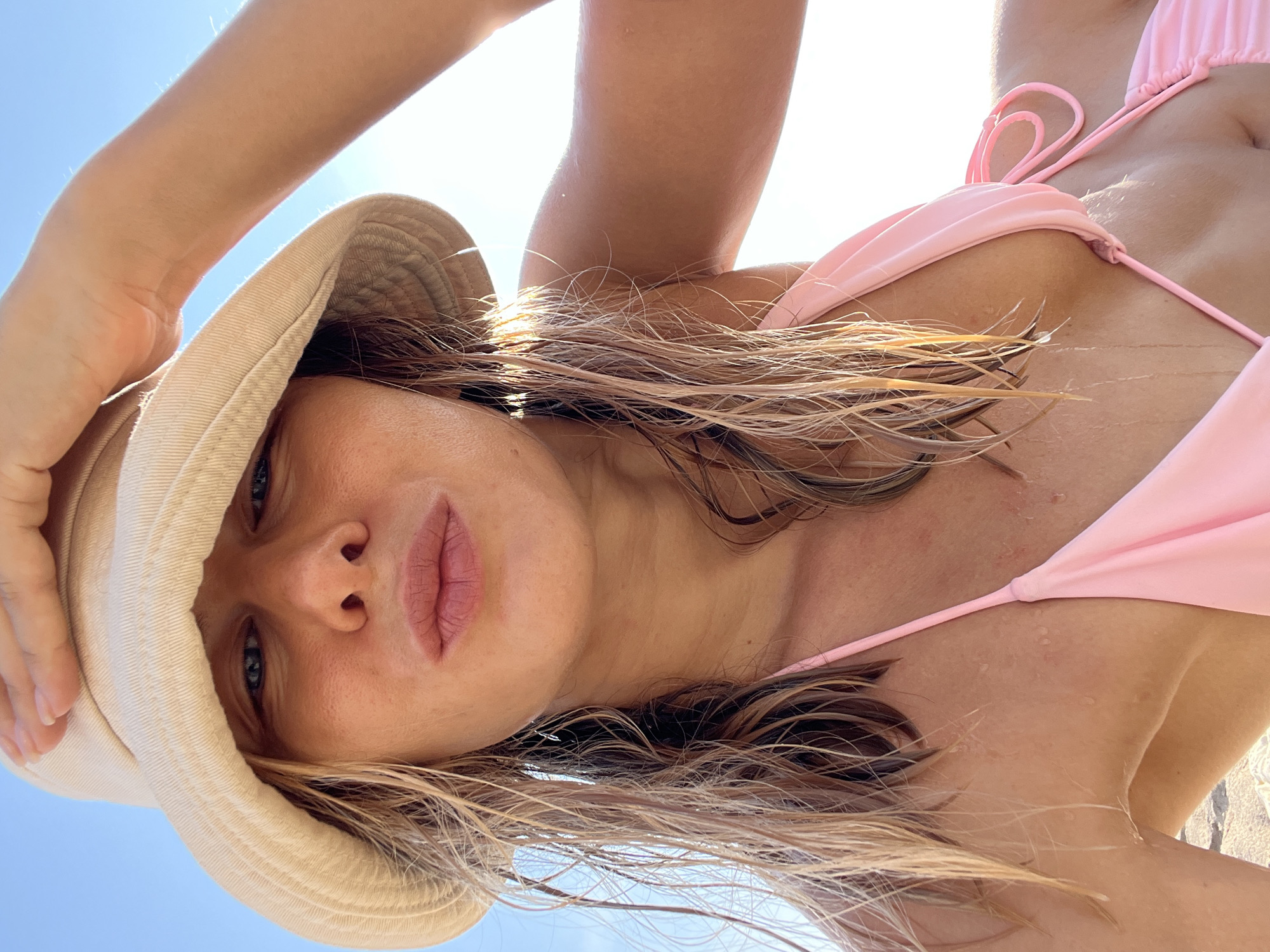 A close-up, low-angle, selfie-style photograph features a person wearing a light-colored bucket hat and a pink bikini top. The individual's right hand is raised to shade their eyes from the bright sunlight, which creates lens flare in the upper right quadrant of the image. The person's hair, which appears wet and light brown, falls around their face and shoulders. The background consists of a clear blue sky. The scene is set outdoors, likely at a beach given the attire and lighting conditions. No specific landmarks of Nha Trang Ward, Vietnam are visible.