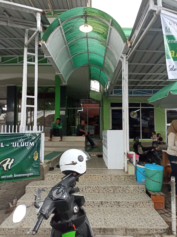 The image shows a scene in front of a building located at latitude: -6.935, longitude: 107.773 in the city of Jatinangor, Indonesia. There is a motorcycle parked in the foreground. The motorcycle is black and has a white helmet on the handlebars. Behind the motorcycle are concrete steps leading to a covered walkway. The walkway is enclosed by a green awning. There are three men sitting on the steps, two on the left and one on the right. On the right is a white cabinet with multiple drawers. There are also two green trash cans on the right. One person is sitting on the ground next to the trash cans. A woman is standing further back. In the background, there is a white and green sign on the right and a green banner with white text on the left. The sign is in Indonesian. The banner is also in Indonesian. The banner reads "IL - ULUM H merah kemenangan". The sky is overcast, and it appears to be raining. There is a white lamp in the center of the awning. The building appears to be a school or a place of worship.