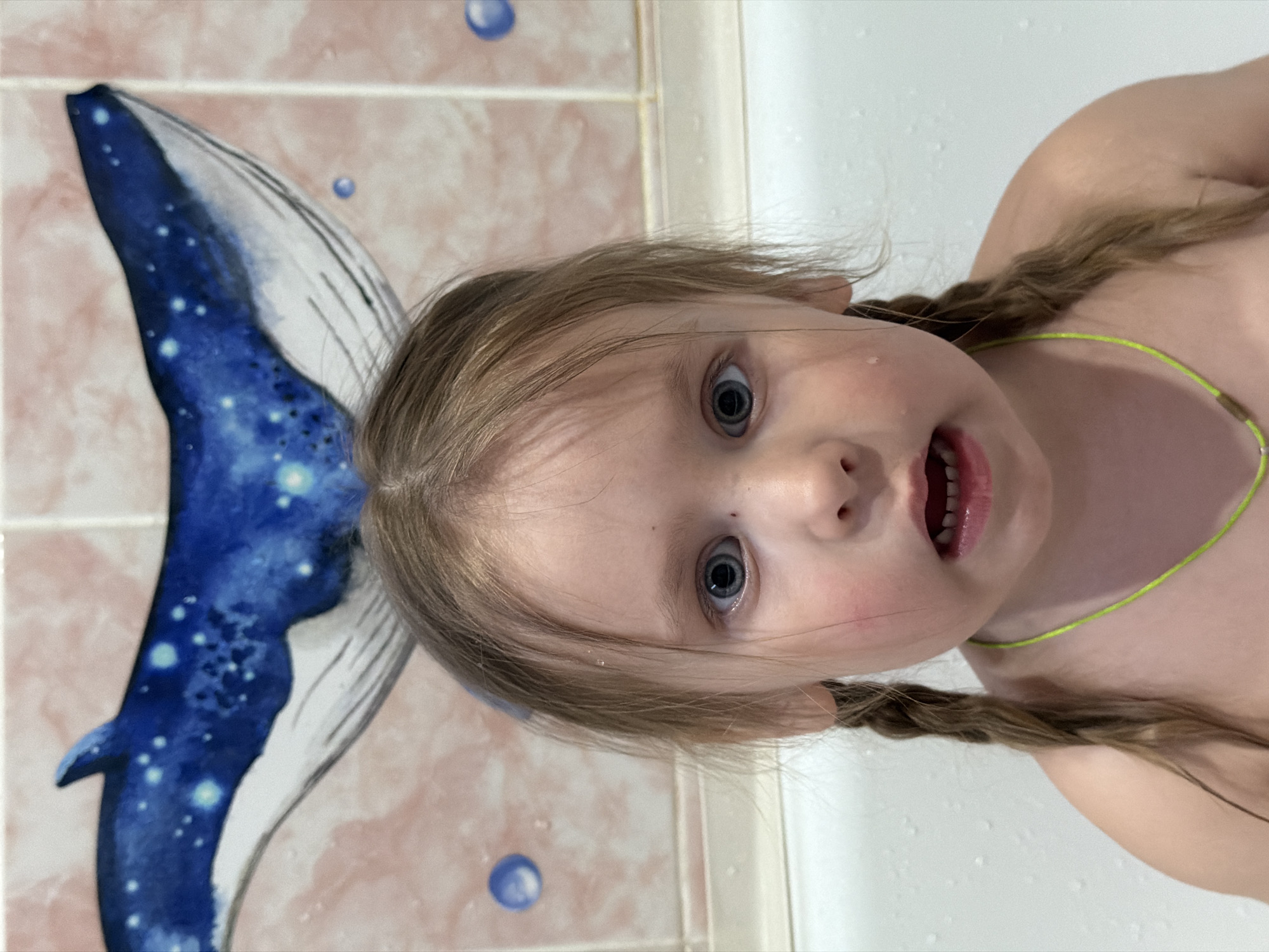 A child, likely a young girl, is photographed from a close-up, low-angle perspective in what appears to be a bathroom. She has light-colored hair styled in braids and is wearing a thin, bright green necklace. She is looking directly at the camera with a slightly open mouth. Behind her, a tiled wall features a painted image of a blue whale tail with white star-like markings. The wall has a pinkish hue with round blue decorations. The background suggests a domestic bathroom setting. There is no definitive information from the image to confirm its location in Chaikovsky, Russia.