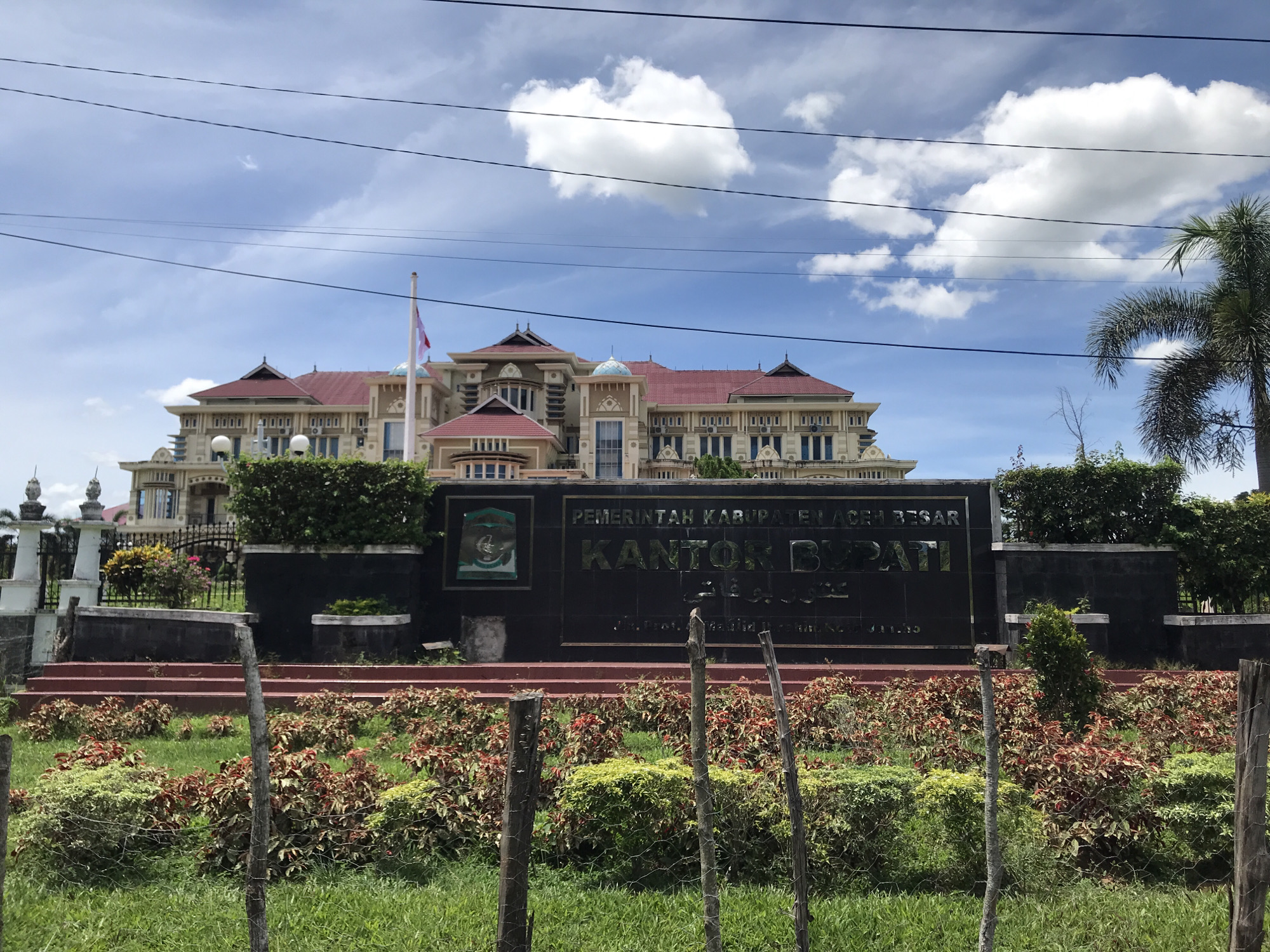 The image shows the Kantor Bupati Aceh Besar (Aceh Besar Regent's Office) in Jantho, Indonesia. The building is a large, multi-story structure with a red tile roof and light-colored facade. A sign in front of the building reads "PEMERINTAH KABUPATEN ACEH BESAR KANTOR BUPATI ACEH BESAR." The sign is black with gold lettering and features the Aceh Besar emblem. The sign is framed by bushes and a low red brick wall. A fence made of wood runs across the front of the image. The sky is blue with white clouds. Power lines run across the sky. A palm tree is visible to the right of the building. A flag is flying from a pole in front of the building.