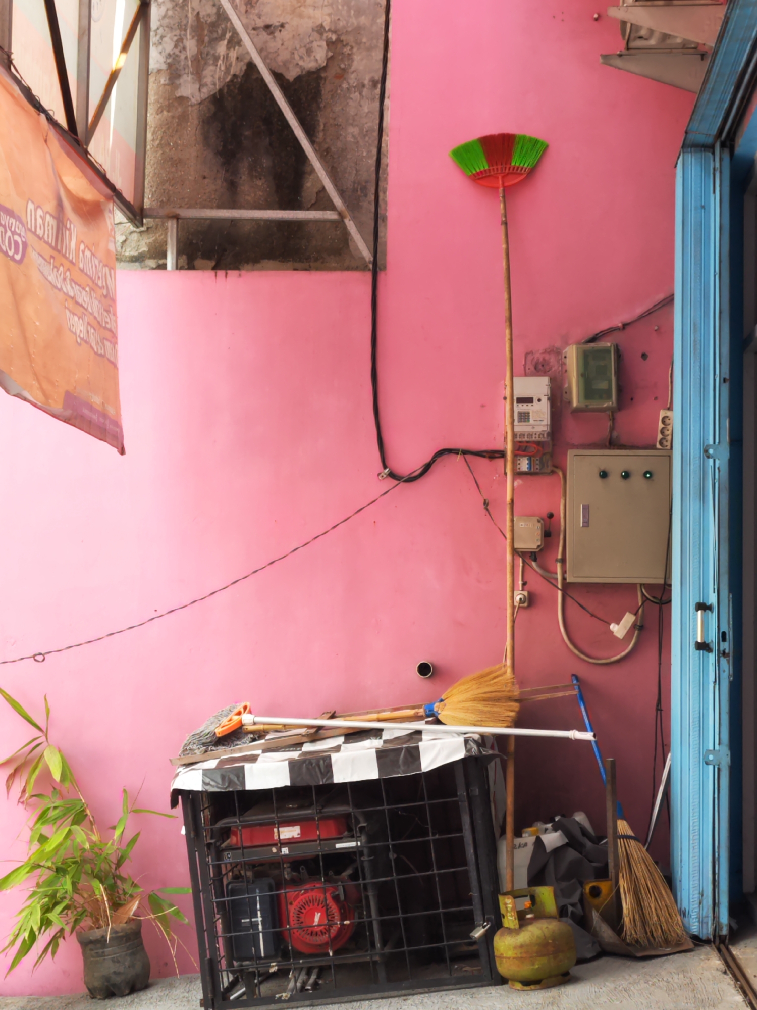 The image depicts an outdoor scene in Sragen, Indonesia, likely a service area or alleyway. The focal point is a vibrant pink wall serving as a backdrop. To the left, an orange banner with text in Indonesian is partially visible, hanging from a metal structure. A potted plant with green bamboo-like leaves sits at the bottom left.
Dominating the center and right side of the image are various utility elements and cleaning tools. A red and green broom stands upright against the pink wall, its bristles fanned out. Below it, several electrical boxes and meters are mounted on the wall, suggesting a utility access point. A generator, covered with a black and white checkered cloth, is visible in the lower center. Two other brooms, one with yellow bristles and another with a blue handle, are leaning against the wall and generator unit. A green gas cylinder is positioned on the ground in front of the generator.
To the far right, a bright blue, corrugated metal door or shutter is partially open, revealing a glimpse of an interior space. The ground appears to be concrete or paved. The overall impression is one of a functional, possibly utilitarian, space with elements of everyday life and maintenance.