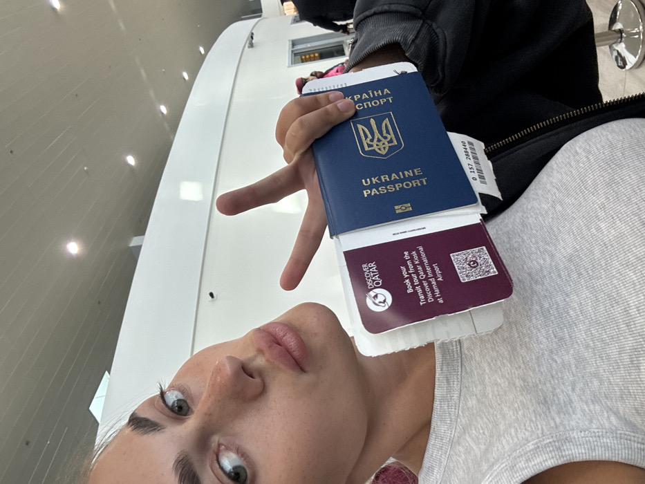 A young adult female is depicted in a close-up, upward-angle selfie within what appears to be a modern airport terminal. She possesses light skin, green eyes, prominent eyebrows, and freckles across her nose and cheeks. Her lips are pursed in a pout, and she looks directly into the camera. She wears a light grey ribbed tank top, with a dark-colored jacket visible over her right shoulder. In her right hand, she holds a blue Ukrainian passport, open to display its cover with the Ukrainian coat of arms and text, along with a "Discover Qatar" leaflet. The leaflet is burgundy, features the "Discover Qatar" logo, and advertises "Book your Transit tour from the Discover Qatar Kiosk at Hamad International Airport." A partial barcode or identification number "0 157 288440" is visible beneath the passport. Her left hand is raised, with the index and middle fingers extended in a 'V' gesture (peace sign). The background consists of a bright, white, modern airport interior with a curved ceiling featuring recessed lighting. Distant, blurred figures are visible further back in the terminal. The specific location in the city of Zanzibar, Tanzania, cannot be confirmed from the visual evidence; the "Discover Qatar" advertisement explicitly points to Hamad International Airport.