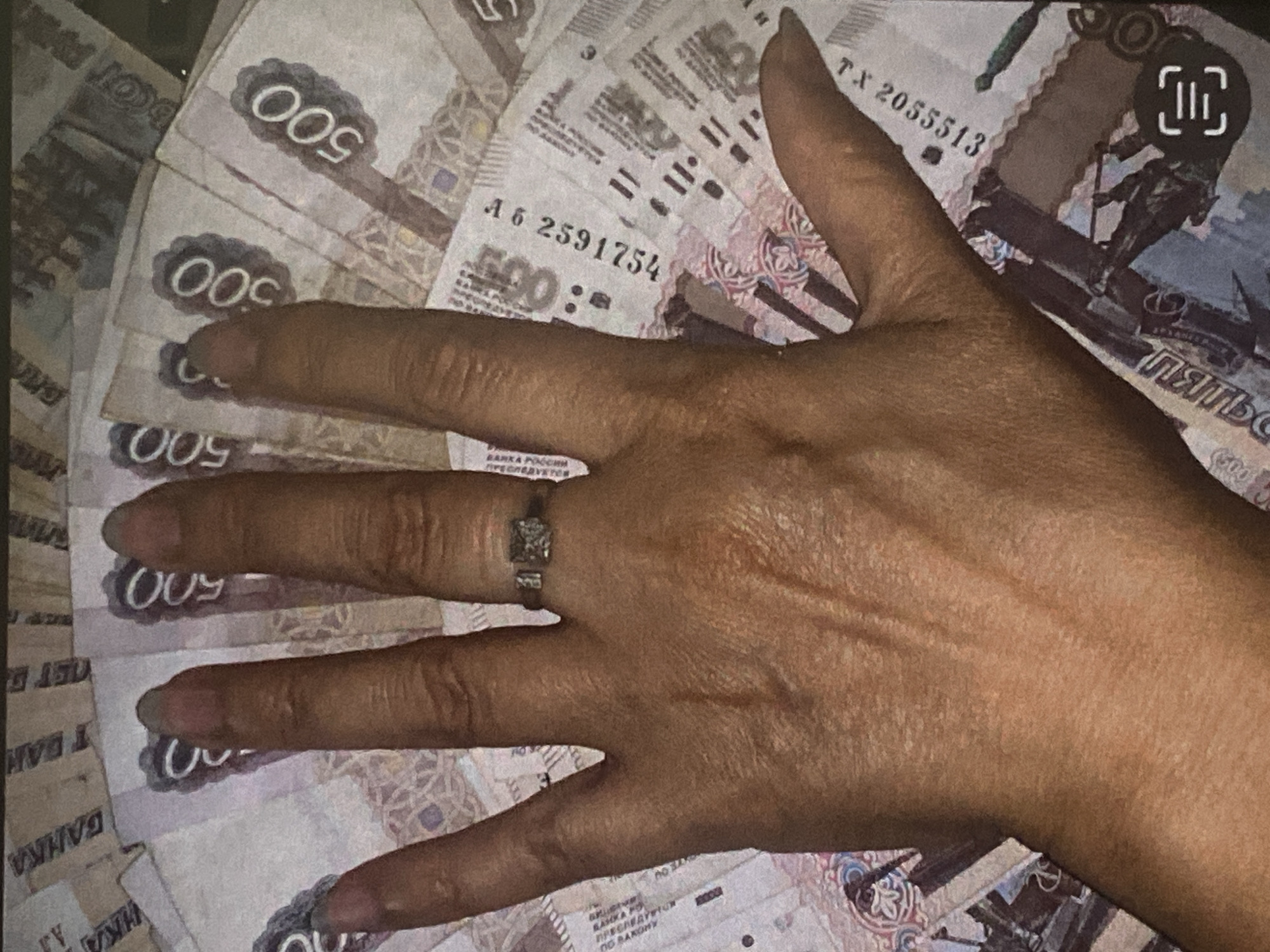 A close-up, overhead shot shows a hand fanned out over a collection of Russian 500 ruble banknotes. The hand is bare except for a ring with a small square-shaped stone on the ring finger. The banknotes are fanned out in a semi-circle, with the numbers "500" visible on several of them. The image appears to be taken indoors, with no specific environmental details visible.