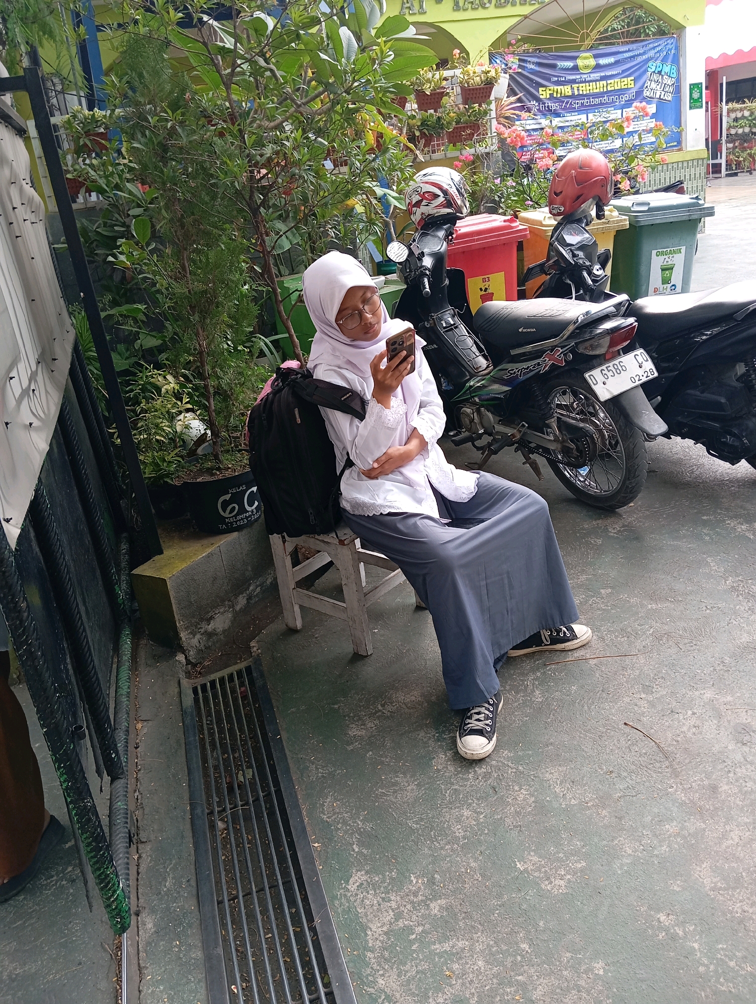 A young girl wearing a white hijab and uniform sits on a white chair next to a black backpack. She is looking at her phone and holding it in her right hand. The girl is sitting in front of several motorcycles and some plants. Behind her, there is a banner that reads "SPMB TAHUN 2026". The scene appears to be in a schoolyard in Bandung, Indonesia, as indicated by the banner which is likely related to university admissions. The ground is paved, and there is a metal grate on the left side of the image. The lighting suggests it is daytime.