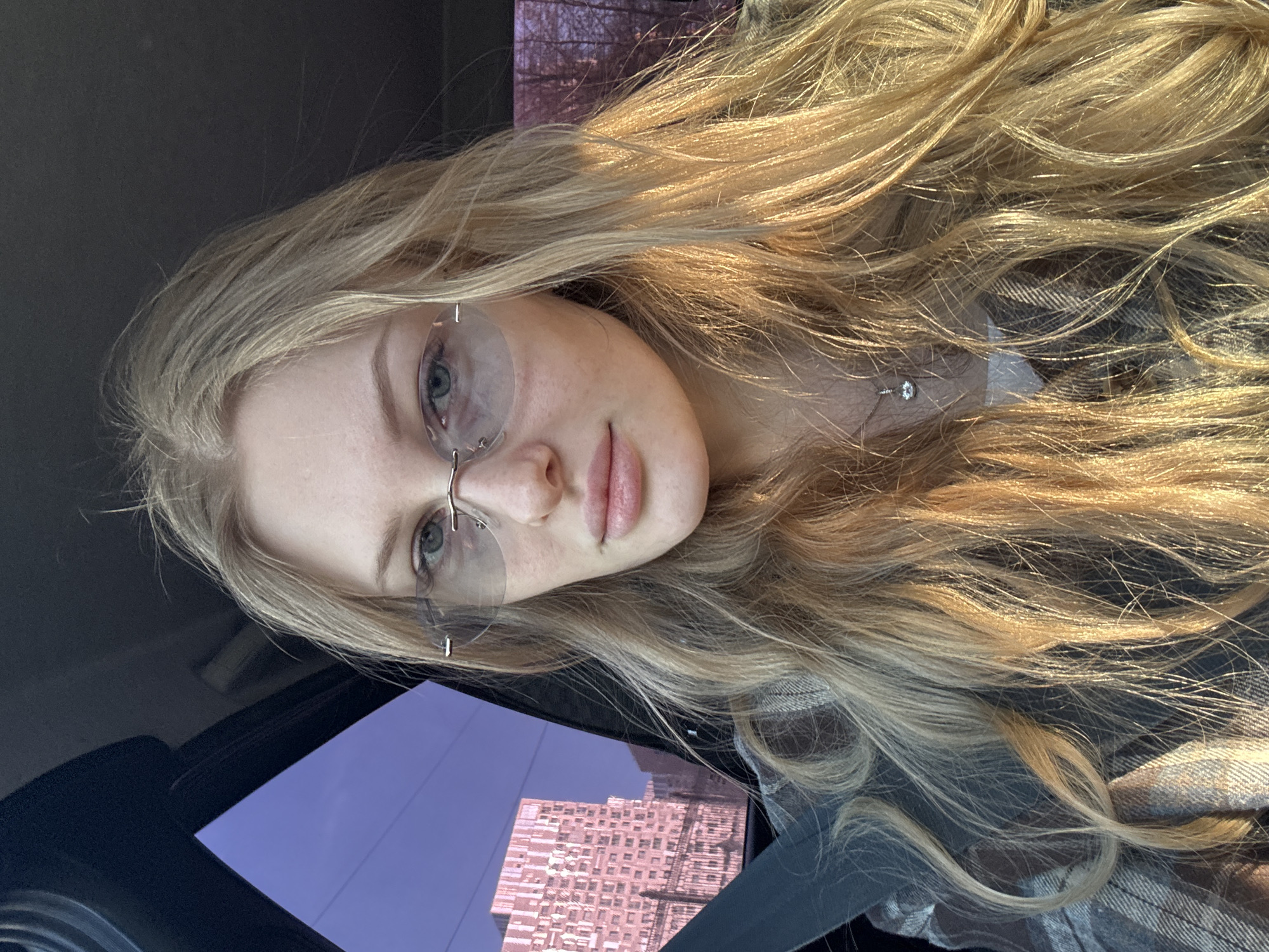 A young, fair-skinned woman with long blonde hair and light-colored eyes poses for a selfie inside a vehicle. She is wearing frameless eyeglasses and a necklace with a small pendant. Part of the dashboard and window are visible, reflecting a pinkish building under a clear blue sky. Based on the architecture of the reflected building and the presence of a vehicle's interior, the location appears to be Perm, Russia. The woman is wearing a plaid shirt. The image has an overall warm lighting.