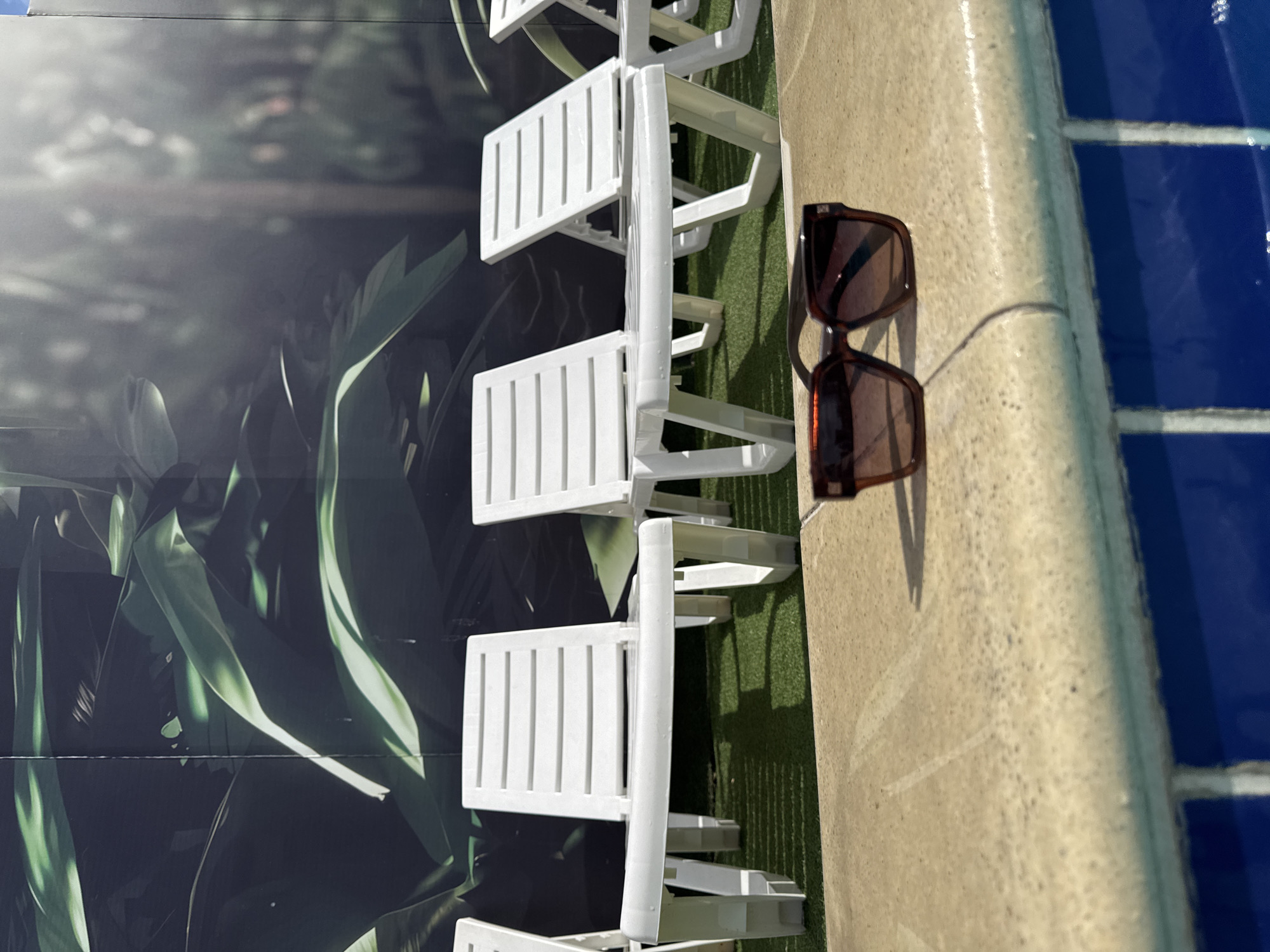 The image captures a bright, sunlit poolside scene, viewed from a slightly elevated angle. In the foreground, a pair of brown-framed sunglasses rests on a light beige, textured concrete or tiled pool deck, casting sharp shadows that indicate strong overhead sunlight.
Adjacent to the concrete deck, a strip of artificial green grass is visible. Lined up on this grass are several white plastic chairs, which appear to be either small-scale models or part of a decorative arrangement, also casting shadows. The chairs feature slatted backs and seats.
To the right, a section of a swimming pool is clearly visible. The water is a vivid blue, with white lines painted on the pool's bottom, and the pool's edge where it meets the deck is gently rounded.
The background consists of a large, dark green mural or backdrop depicting lush foliage, with prominent leaves and highlights suggesting depth and a natural, verdant environment. This backdrop creates the impression of a tropical or garden setting.
The scene suggests a warm, sunny daytime setting, likely an indoor pool area or a creatively designed outdoor space, intended for relaxation and leisure. No people or visible text are present in the image. The overall impression is one of tranquility and leisure.