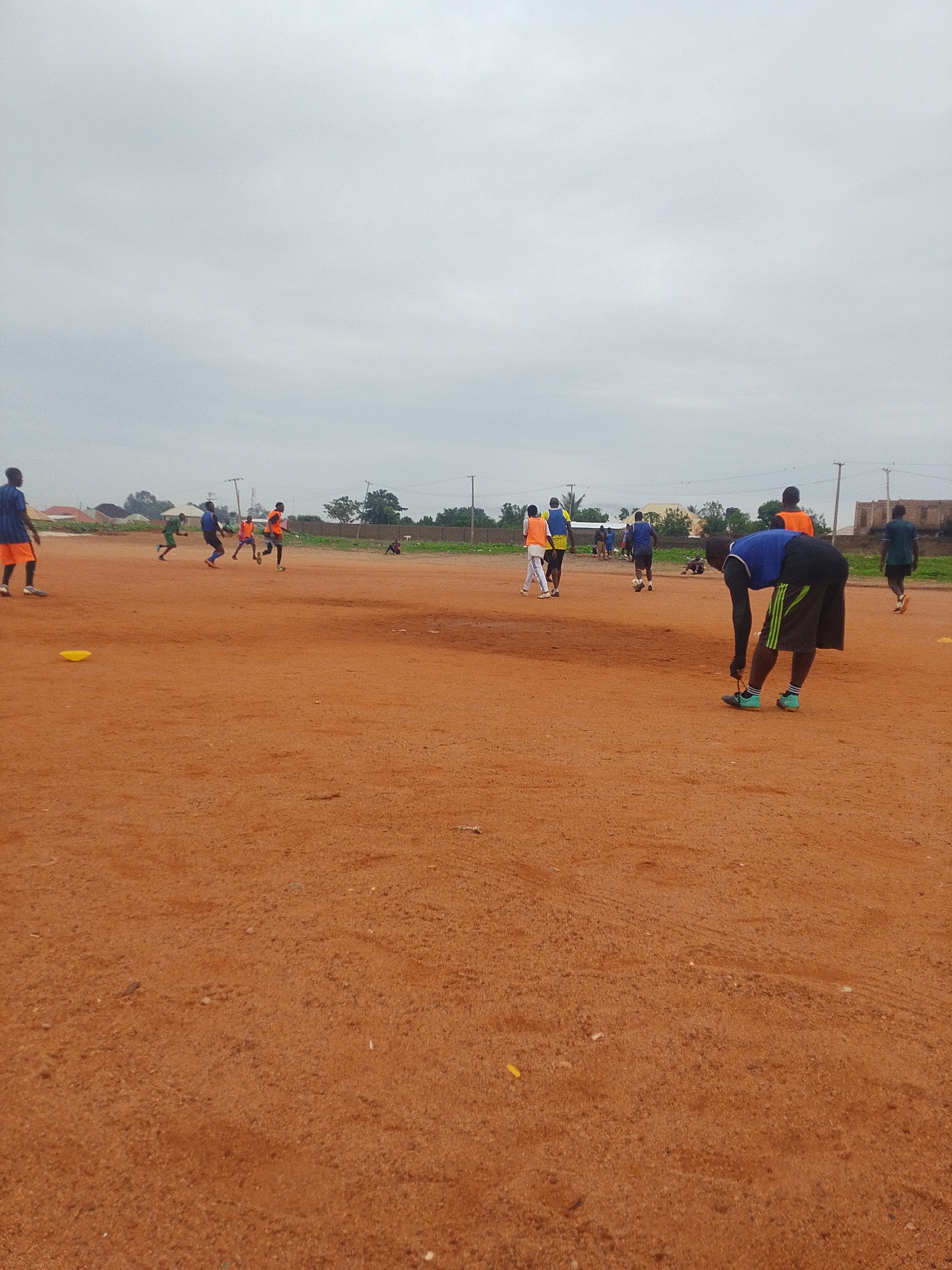 The image depicts a soccer field with a reddish-brown dirt surface. Several people, presumably soccer players, are present on the field. Some are actively playing soccer, while others are standing or bending over. The players are wearing a variety of athletic clothing, including blue, orange, green, and black. A yellow cone sits on the ground in the foreground. The background shows buildings and trees, suggesting the field is located in a somewhat urban or suburban area. The sky is overcast. The location is Unknown, Unknown.