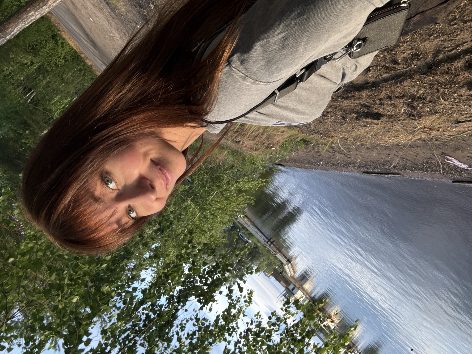 The image is a close-up, low-angle selfie of a young woman with long, reddish-brown hair and green eyes. She is wearing a grey hooded sweatshirt and a black crossbody bag. Her head is tilted, and she is looking at the camera with a slight smile. Sunlight is filtering through the trees, suggesting it is daytime. The background shows a body of water, possibly a river or lake, with some structures and trees along the shore. The overall scene appears to be outdoors, in a natural setting. The orientation of the image is portrait, with the subjects appearing sideways. The location is identified as Chaikovsky, Russia.