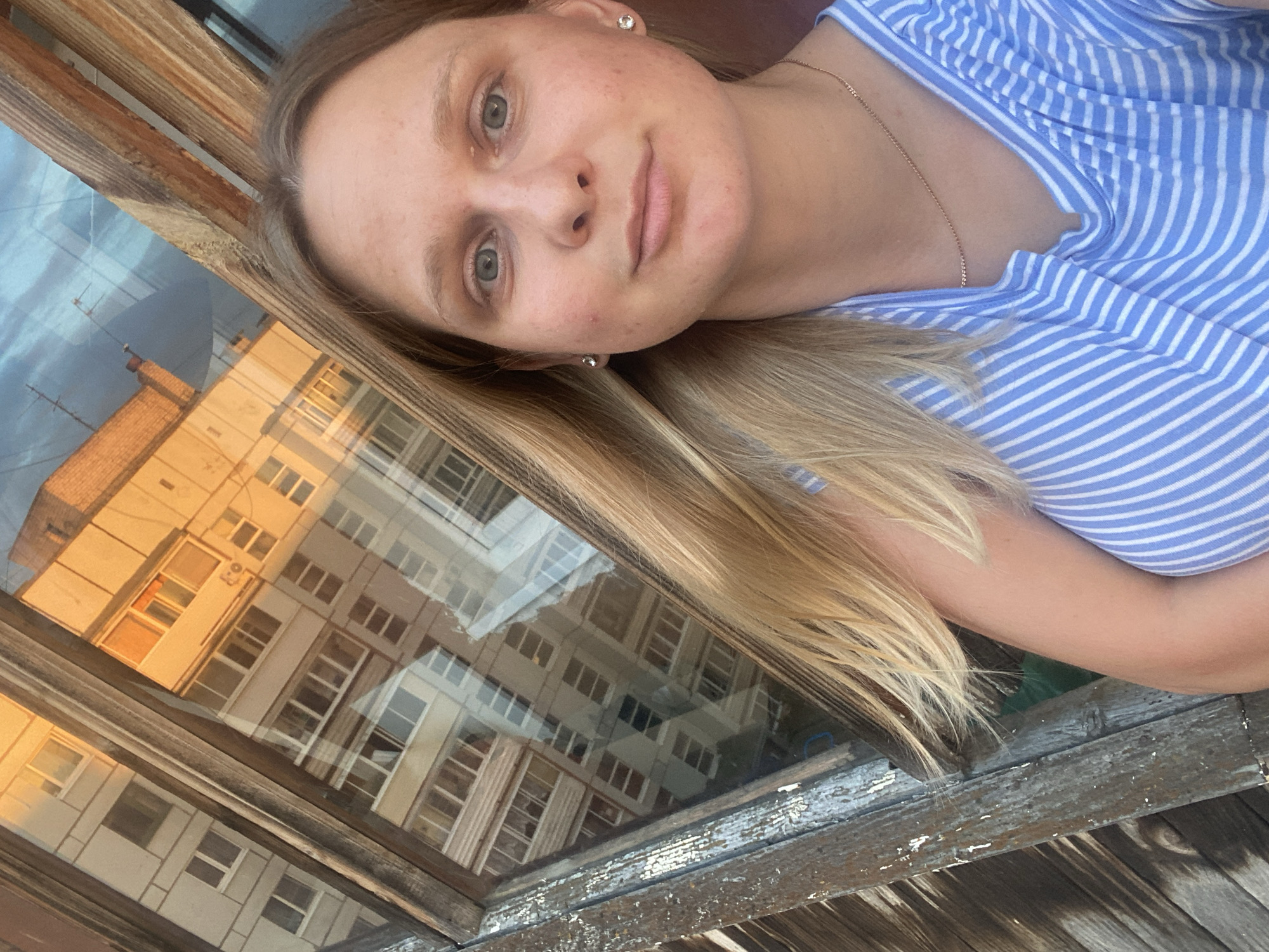 A young Caucasian woman with long blonde hair and fair skin takes a selfie. She is wearing a blue and white striped t-shirt and a delicate gold chain. She has light blue eyes and minimal makeup, with some visible blemishes on her face. She is positioned in front of a weathered wooden window frame. The windowpane reflects a multi-story apartment building bathed in the warm glow of what appears to be sunset or sunrise. The building's architecture suggests it is located in a typical Soviet-era residential area. The specific location is the city of Chaikovsky, Russia, identifiable by the building style and the overall urban environment. The scene captures a moment of casual self-photography, with the backdrop of urban architecture and natural light.