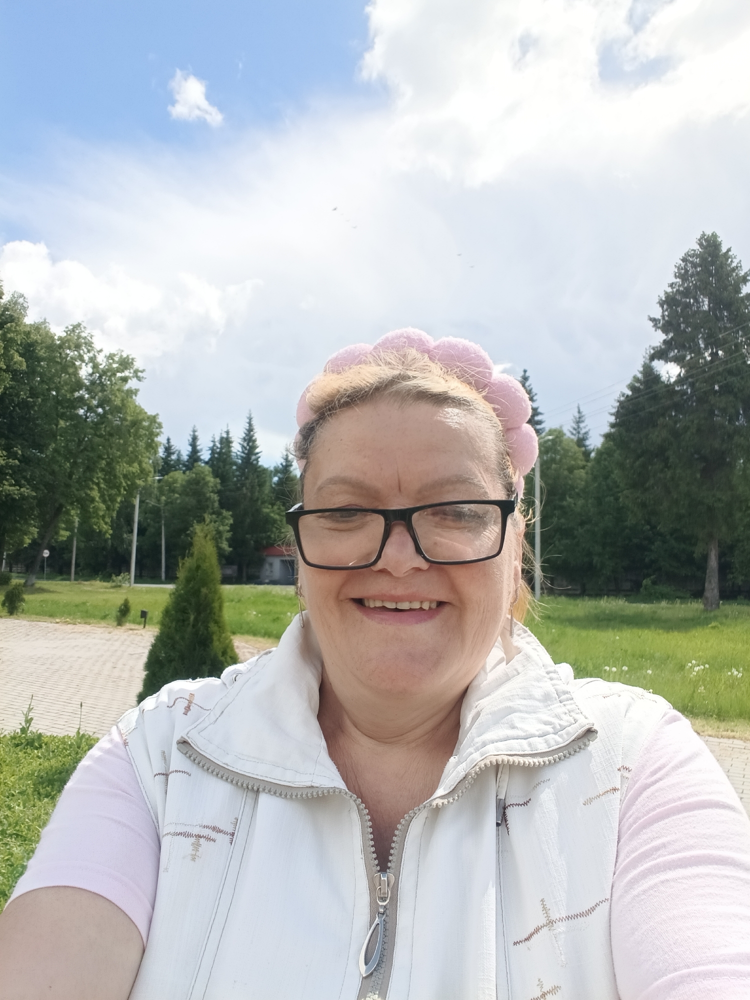 A woman with blonde hair wearing glasses and a pink headband with pom-poms takes a selfie outdoors. She is wearing a pink t-shirt underneath a white vest. Behind her, there is a paved area and green grass. In the background, there are coniferous and deciduous trees, some of which are shaped like cones. The sky is blue with white clouds. This location is likely Красный Маяк, Russia.