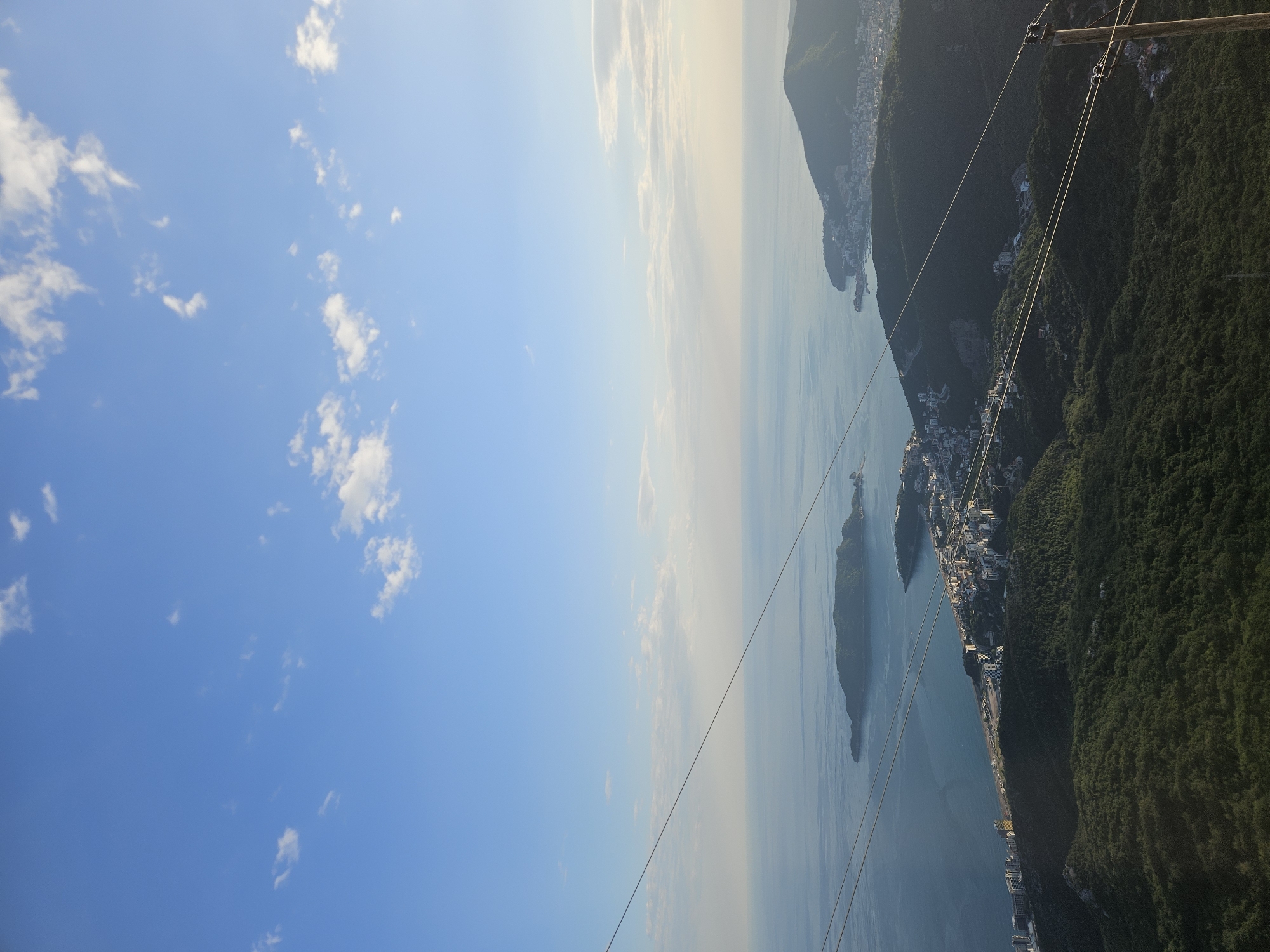 The image presents an aerial view featuring the coastal landscape of Brajici, Montenegro. Predominantly blue sky with scattered clouds occupies a significant portion of the left side. Moving towards the right, a large body of water is visible, possibly the Adriatic Sea, with a couple of islands. A coastline lined with buildings can be seen, with dense, green mountain terrain to the right. Overhead cable lines cut diagonally across the scene.