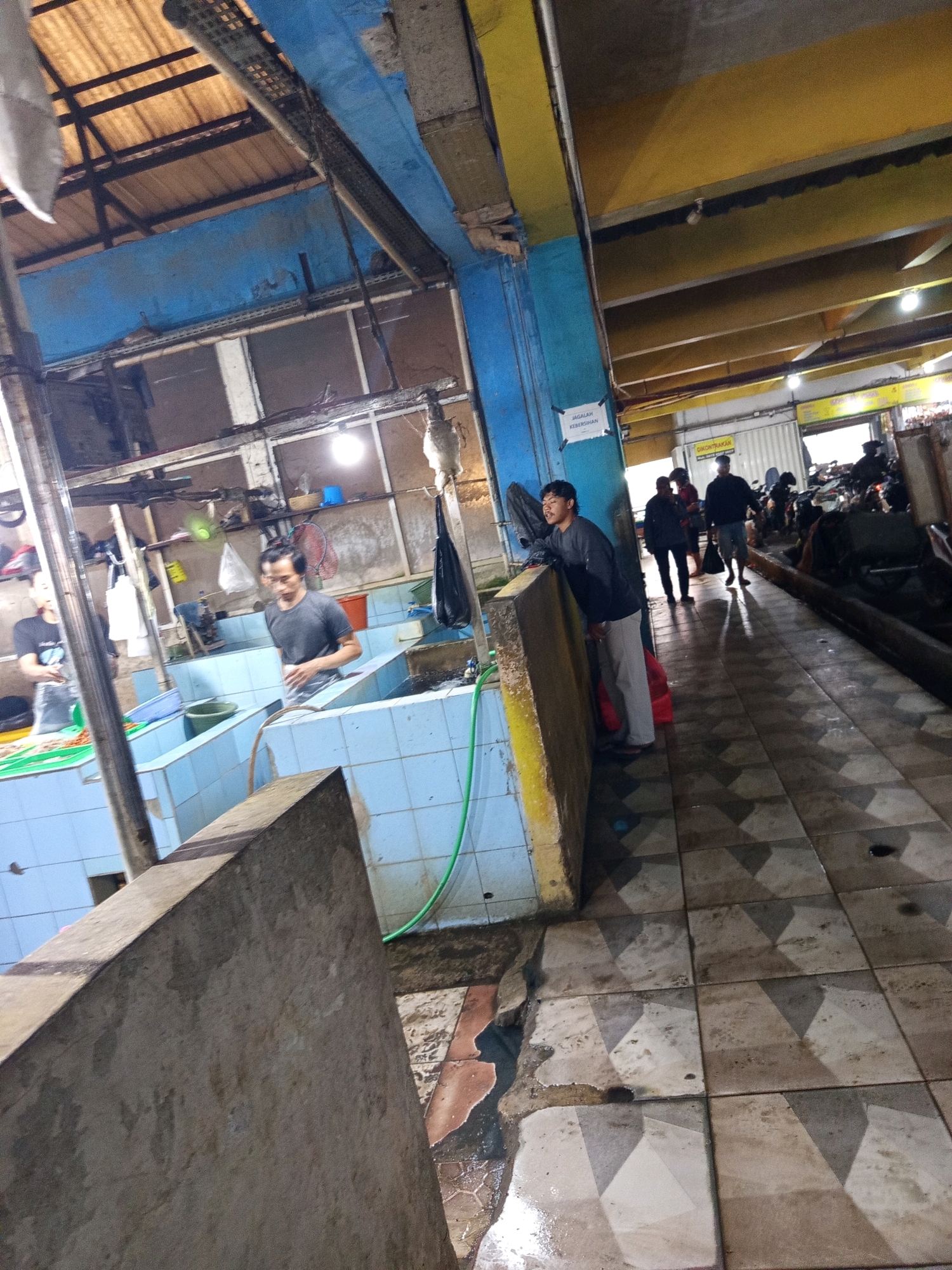 The image depicts an indoor market or a fish processing facility in Karawang, Indonesia. In the foreground, a person wearing a dark t-shirt and light-colored pants is leaning against a yellow and concrete partition. To the left, a man in a grey t-shirt is standing in a tiled pool, seemingly working. Another person is partially visible in the background, also near a pool. The environment is utilitarian, with blue tiled walls and a tiled floor that appears wet. Overhead, the ceiling shows exposed beams and a corrugated roof. In the background, several motorcycles are parked, and other individuals are visible, suggesting a bustling commercial area. A sign with Indonesian text is present on a blue pillar. The lighting appears to be artificial, with a bright light bulb illuminating a portion of the space.