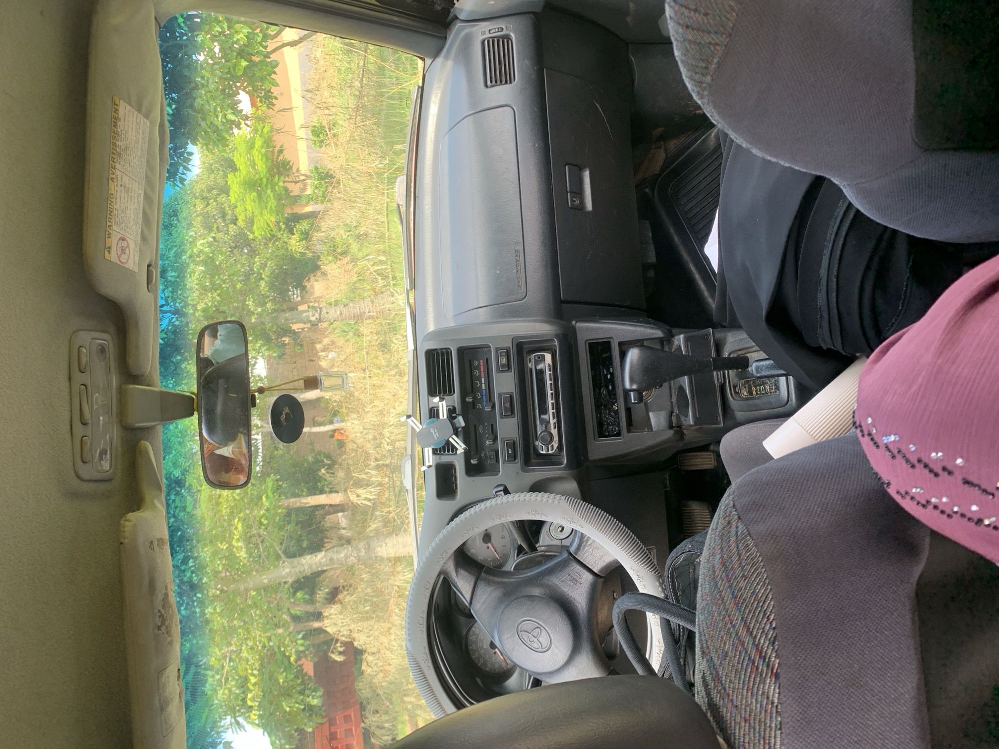 The image shows the interior of a car, likely parked outdoors in a rural or semi-rural area. The foreground is dominated by the seats of the car, with a person wearing a pink garment visible in the bottom right. The dashboard is visible, featuring a steering wheel with a grey cover, instrument cluster, and a radio. A car phone holder is attached to the air vent. The exterior environment, visible through the windshield, consists of trees and dry vegetation. A red structure, possibly a truck or building, is partially visible to the left. The lighting suggests it is daytime, with sunlight filtering through the trees.