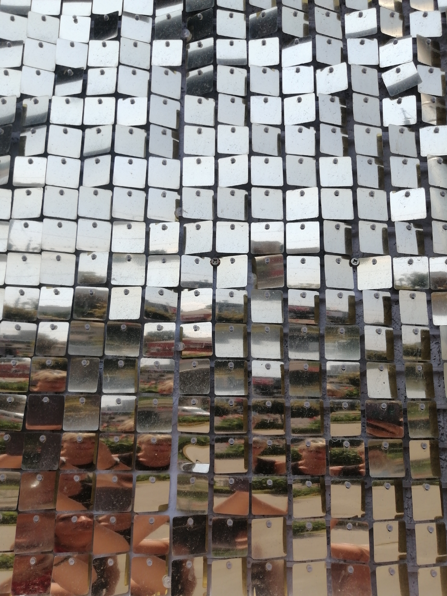 The image is a close-up, high-angle, full-frame shot of a wall covered in square, reflective, silver sequins. The sequins are arranged in a grid pattern, with each square tilted slightly, creating a shimmering effect. The sequins are attached to a dark backing material, and many have small circular holes in the center, suggesting they are either sewn or pinned on. The surface of the sequins reflects the surrounding environment, showing distorted images of what appears to be an outdoor scene with greenery and possibly people. The lighting is bright and even, highlighting the reflective quality of the sequins. There is no discernible location in the city of Kremenchuk, Ukraine, visible in the image.