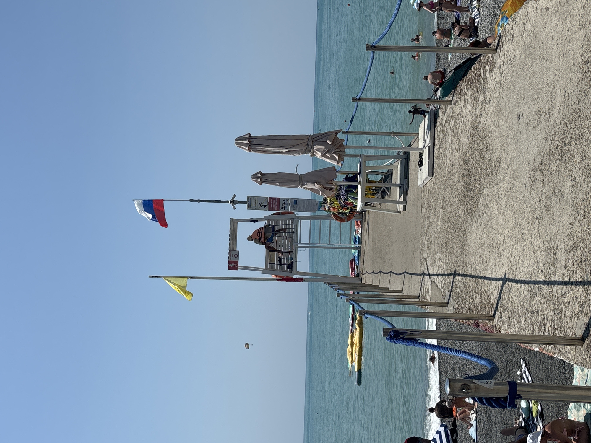 This image depicts a sunny day at a beach in Sochi, Russia. A concrete pier extends into the turquoise sea, lined with a blue rope railing. On the pier, a lifeguard tower stands prominently, with a lifeguard seated at the top wearing an orange vest. Two flags, one yellow and one Russian tricolor, fly from poles near the tower. Several closed beach umbrellas are arranged on a white table. In the distance, people are swimming and relaxing on the pebble beach. A parasail is visible in the sky. The overall scene conveys a sense of a typical summer day at a seaside resort.