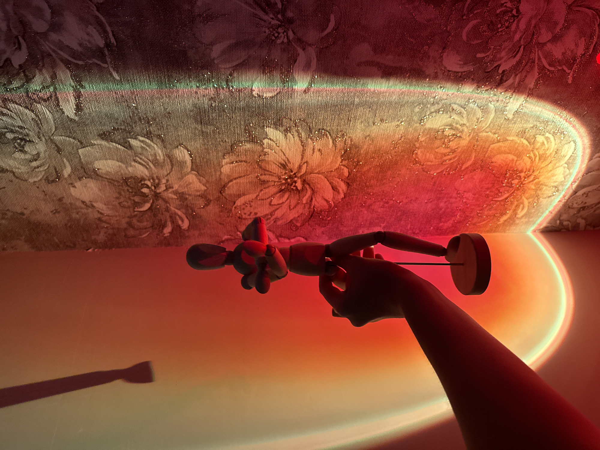 This media file depicts a dimly lit room with a floral patterned wallpaper. A human hand is holding a wooden mannequin. The room is illuminated by a projected sunset light, casting a gradient of red, orange, and yellow hues across the wall and floor. The light creates a soft, glowing effect, highlighting the textures of the wallpaper and the wooden mannequin. The mannequin appears to be in a neutral pose, held by the hand as if it's being displayed or manipulated. The overall mood is artistic and atmospheric, with the warm lighting creating a sense of intimacy and wonder. The time of day is suggested to be evening or night due to the artificial sunset lighting. The location is indoors, as indicated by the wallpaper and the controlled lighting. There is no visible text in the media file.