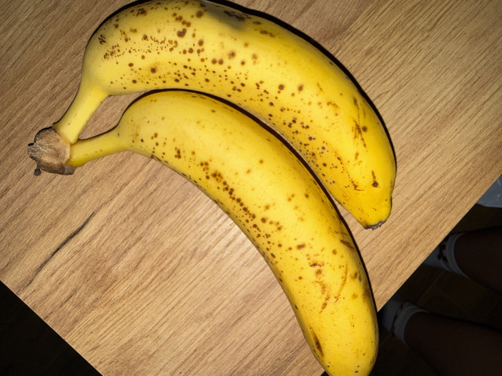 Two ripe bananas with brown spots lie on a wooden surface. The bananas are yellow with a slightly curved shape. The wooden surface has a visible grain pattern and is light brown in color. The image is taken from a top-down perspective. There are no people or animals visible in the image. The location in Varna, Bulgaria cannot be determined from the image.
