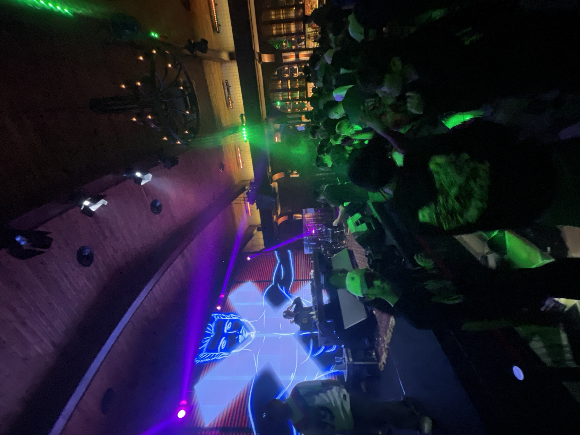 The image depicts a nighttime event in North Jakarta, Indonesia, characterized by a lively atmosphere with a DJ performing on a stage. The DJ is seen behind a console, illuminated by bright purple and blue lights, with a large digital screen displaying a stylized animated figure as a backdrop. A crowd of people is gathered in front of the stage, some dancing and interacting. Green laser lights are also visible, cutting through the darkness and adding to the dynamic ambiance of the venue. The ceiling of the venue features a large, ornate chandelier and exposed wooden beams, suggesting a stylish and possibly upscale establishment. The overall scene suggests a concert, club, or music festival.