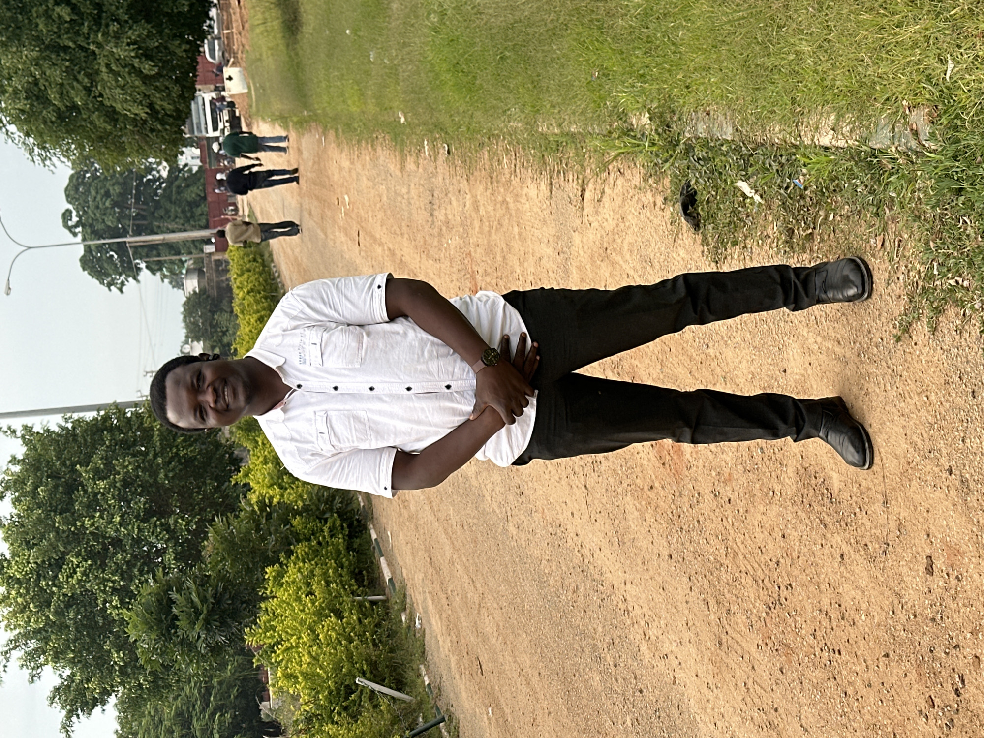 A full-body, medium shot depicts a man standing on a dirt road in Dadi Riba, Nigeria. The man, who appears to be in his late 30s or early 40s, is wearing a white short-sleeved button-down shirt, dark trousers, and black shoes. He has his hands clasped in front of him. He is smiling and looking directly at the camera.
The road is unpaved and appears to be made of dirt and gravel. On the left side of the road, there are dense green bushes. On the right side of the road, there is a patch of green grass. In the background, there are several trees, a streetlamp, and some buildings. There are also several other people walking on the road in the distance, as well as a truck. The sky is overcast.