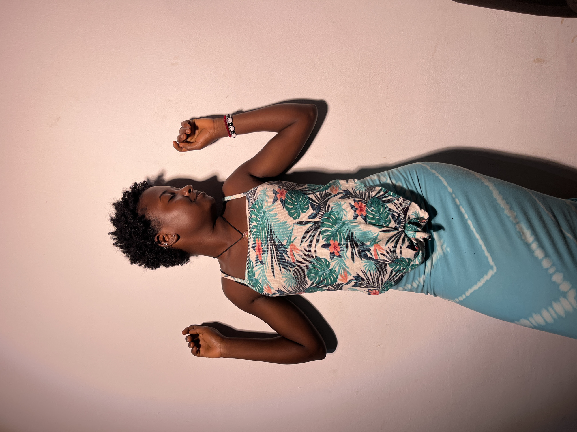 A young Black woman with short, dark, curly hair stands with her eyes closed and head tilted down, her face in profile. She is wearing a white tank top with a tropical floral print of green leaves and coral-colored flowers, and a tied knot at the front. Her lower body is clad in a light blue, tie-dye patterned maxi skirt. Her arms are raised to shoulder height, bent at the elbows, with her fists clenched. She has a thin necklace around her neck and a multi-colored bracelet on her right wrist. The woman is positioned against a plain, pale pinkish-white wall, with a noticeable shadow cast on the wall behind her and to her left. The lighting appears to be coming from the right, creating a dramatic effect with strong shadows. The overall setting is indoors, with no visible elements of an urban environment.