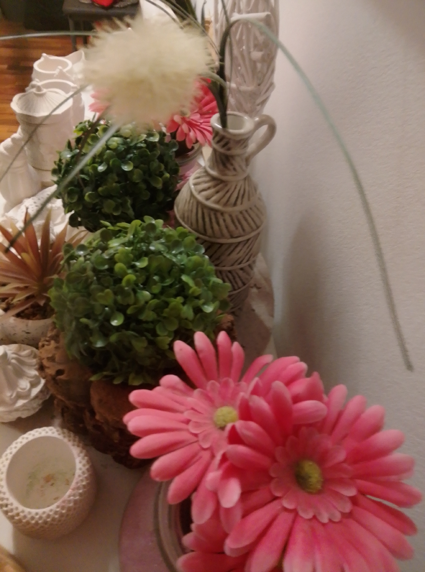 The image displays a close-up view of a decorative arrangement of artificial plants and objects, likely an indoor display. The arrangement includes two green, spherical artificial plants, a light brown artificial succulent, a cream-colored vase with a textured, rope-like design, a fluffy white artificial flower, and two bright pink artificial daisies in the foreground. There are also several white decorative ceramic pieces visible in the background. The items are arranged on a flat surface, possibly a shelf or table, against a light-colored wall. There is no discernible indication of the city of Wedel, Germany, or any specific location within it, in this image.