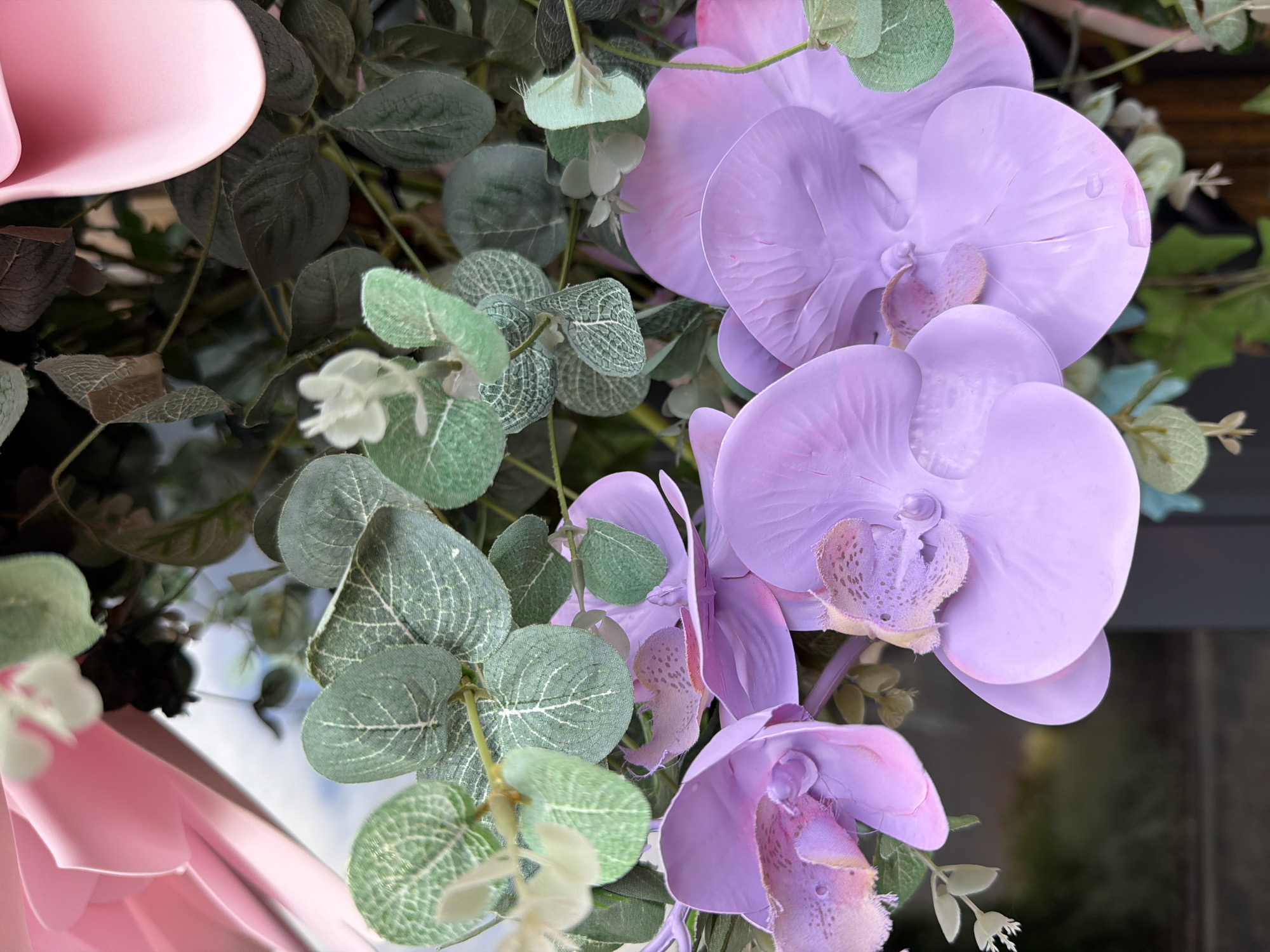 The image shows a close-up of artificial purple orchids with green eucalyptus leaves. A pink ribbon is visible in the upper left corner. The background is blurred, suggesting an indoor setting with some light coming from outside. There are no people or discernible actions taking place. The image is a still life of decorative floral elements.