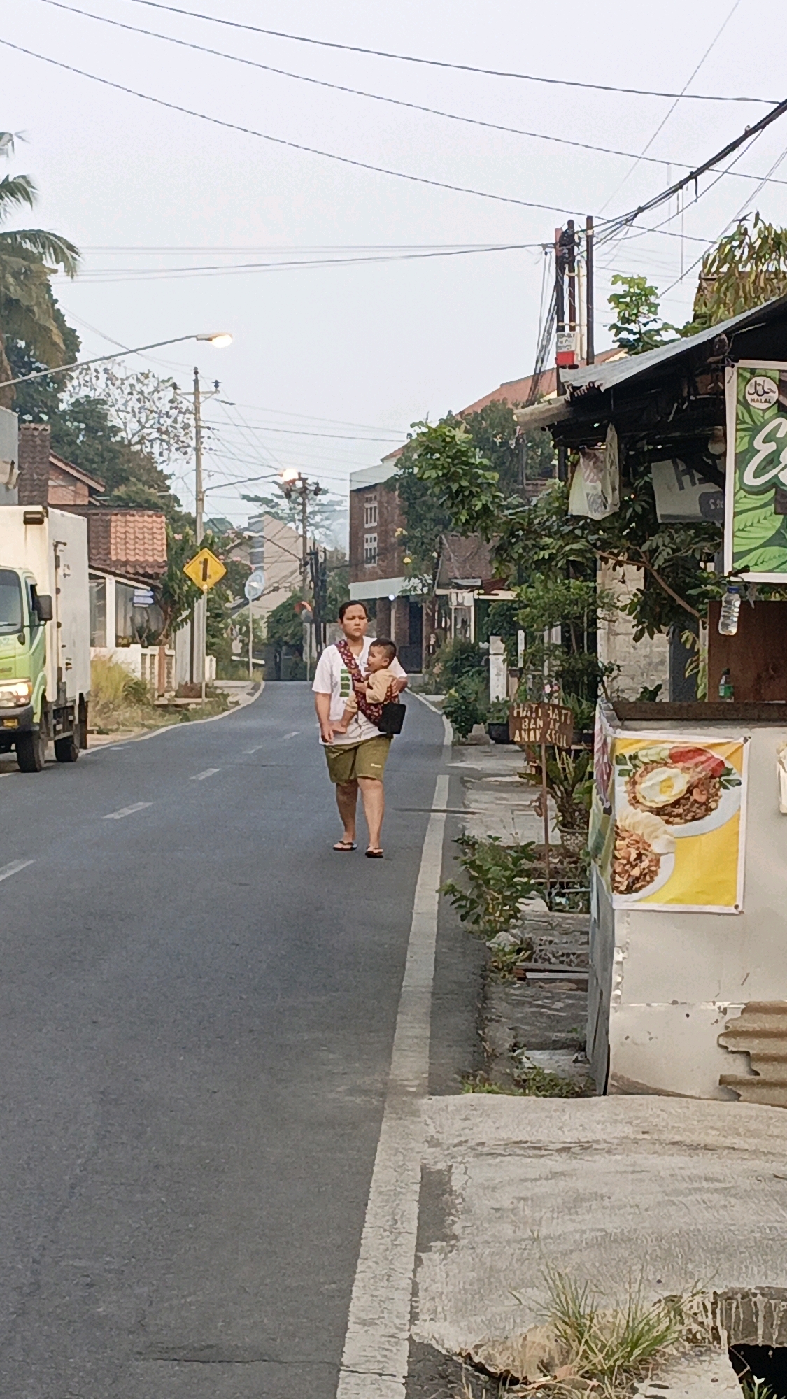 A woman walks on the side of a street in Salatiga, Indonesia, carrying a baby in a sling. The woman is wearing a white t-shirt and khaki shorts. The baby is wearing a light brown outfit. They are walking on the right side of a two-lane asphalt road, with a white line marking the edge of the road. On the left side of the road, there is a white truck. Buildings and trees line the right side of the street, with a few signs visible. The sky is overcast.