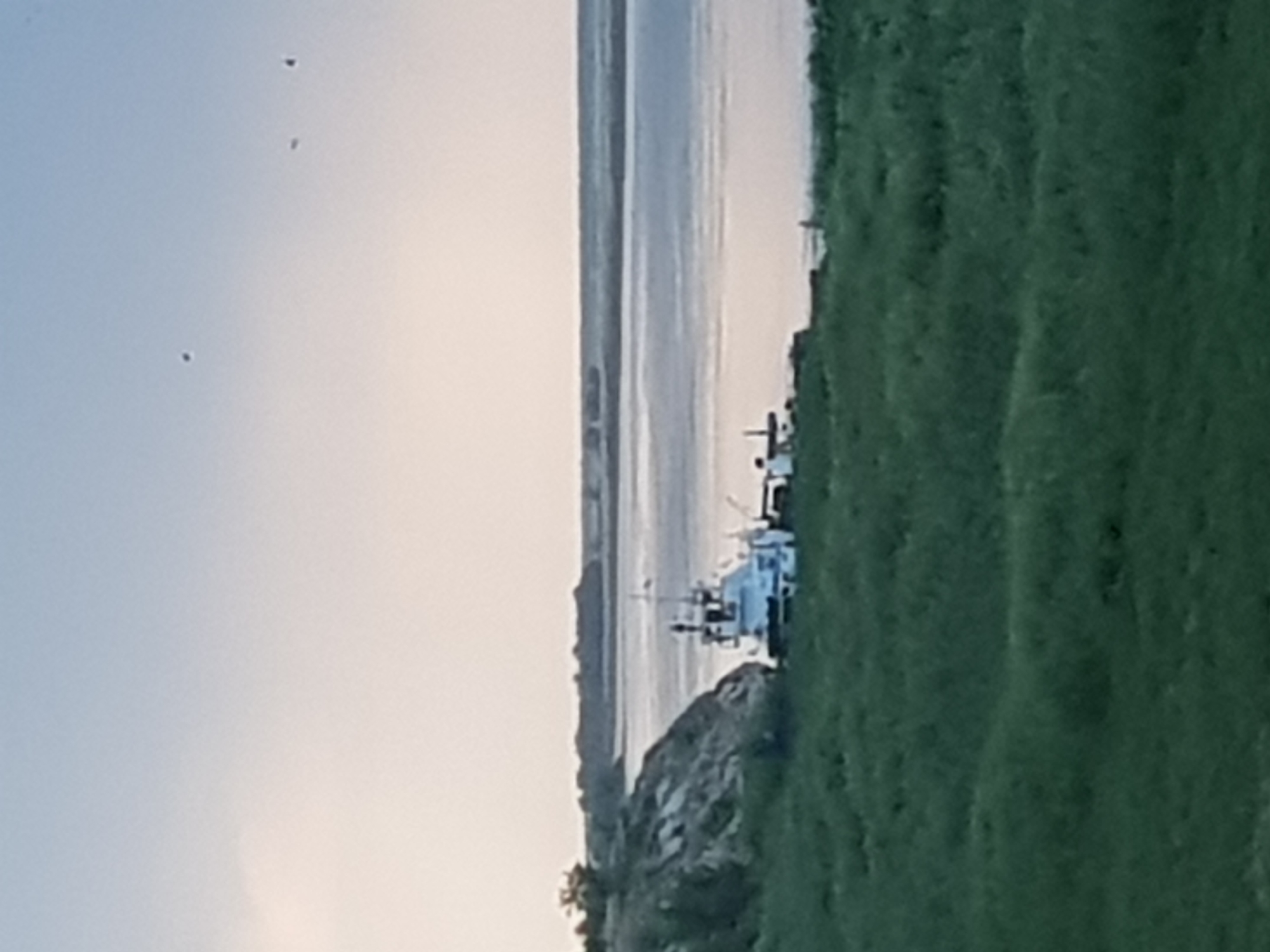 A tugboat is docked near a rocky incline covered with grass and trees, beside a wide body of water. The water stretches to the horizon, where it meets a hazy, muted sky. In the foreground, a grassy slope descends towards the water. Several small, dark specks, possibly birds, are visible in the upper portion of the sky. The scene suggests a calm, natural setting, likely near the Irtysh River, which flows through Kurchatov, Kazakhstan.