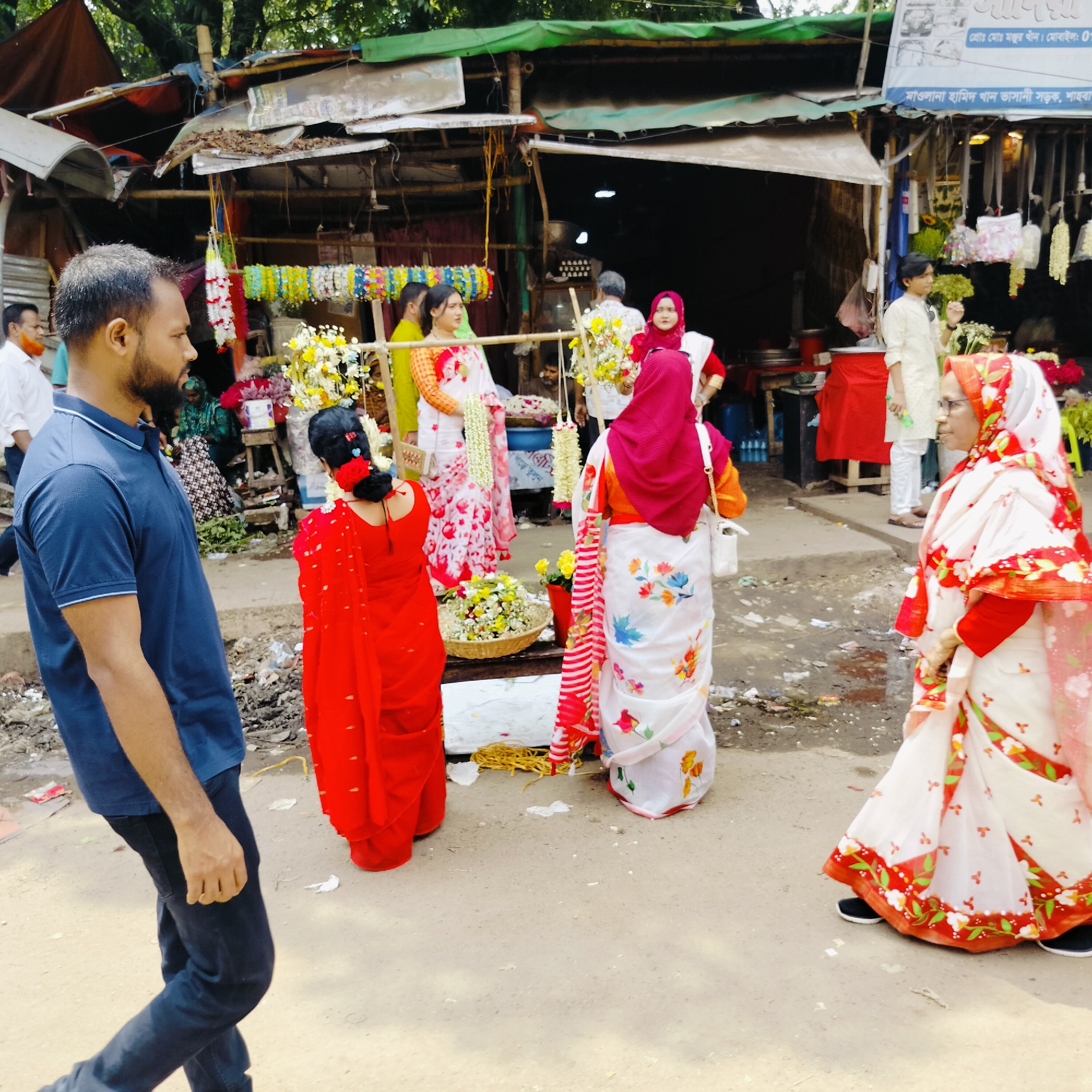 This medium shot photograph captures a bustling street scene in Dhaka, Bangladesh, identified by the sign "মাওলানা হামিদ খান ভাসানী সড়ক, শাহবা" indicating the road Maulana Hamid Khan Bhasani Road in Shahbag. A man with a beard, clad in a blue polo shirt and black jeans, is walking along the road from left to right. Several women are gathered in the middle ground, adorned in colorful saris and potentially engaged in commercial activities. One woman is dressed in a vibrant red sari. Another one is wearing a white sari with red and floral prints, and a red headscarf. A third woman is wearing a white sari with floral prints, and a red and white striped stole. A basket of flowers is placed on a table, suggesting they might be selling them. In the background, there's a makeshift shop selling garlands and flowers, typical of street vendors in the area. Other individuals are milling about near the shop, and a couple of men wearing more traditional garb can be seen as well. The setting appears to be a busy, open-air market or commercial area. The ground is somewhat uneven and appears littered.