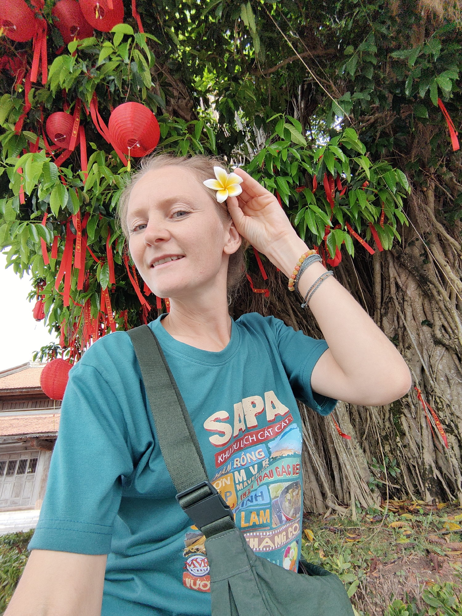 A woman poses for a selfie outdoors, standing in front of a large tree adorned with red lanterns and ribbons. She is holding a white and yellow flower near her ear. The woman is wearing a teal t-shirt with an image and the word "SAPA" on it, along with an olive green shoulder bag. She also has bracelets on her wrist. The background reveals part of a building with a traditional roofline. The scene is set in Hong Gai Ward, Vietnam, as indicated by the text on the shirt.