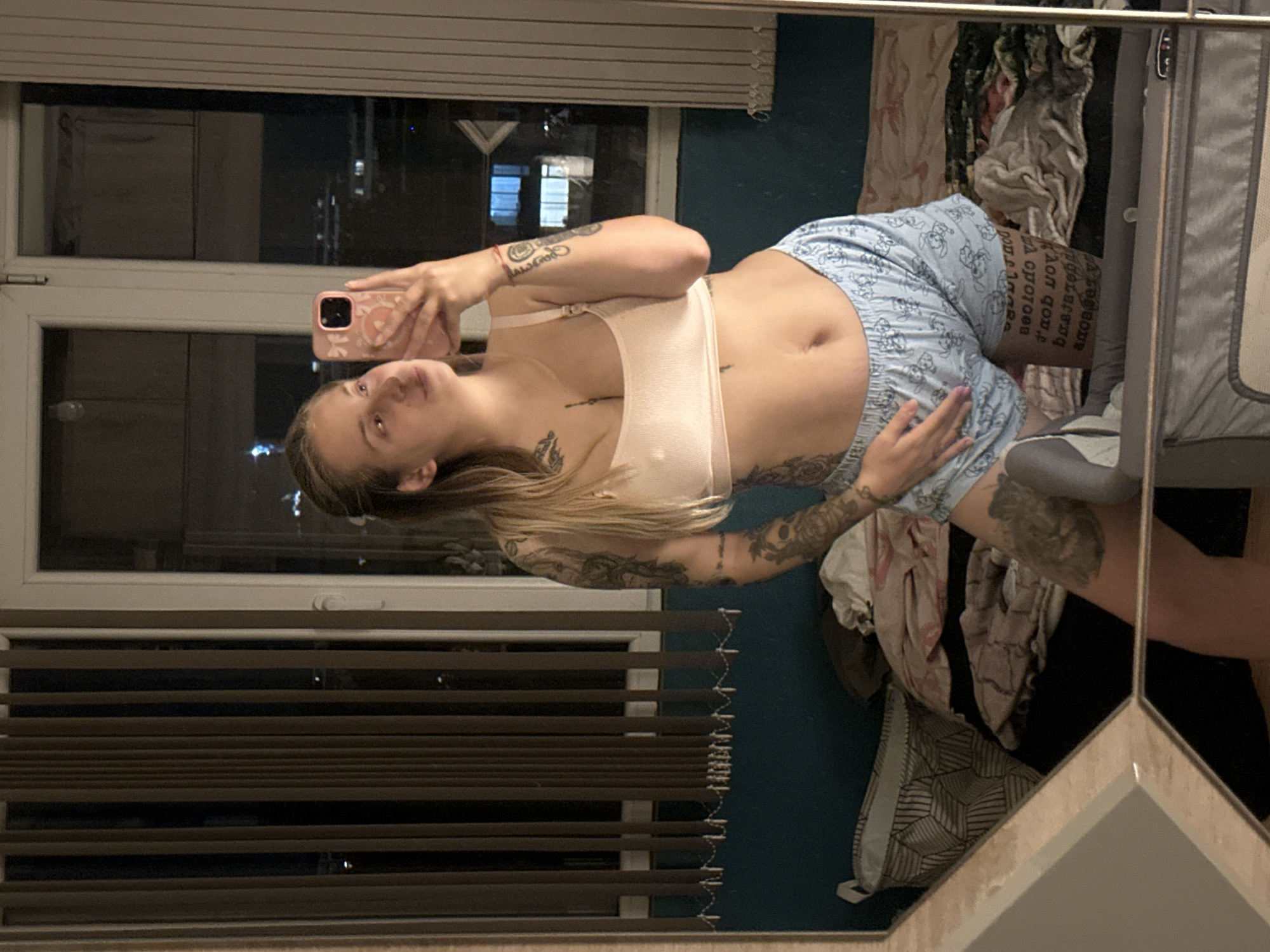 A young woman with blonde hair pulled back and visible tattoos on her arms and legs is taking a mirror selfie in a bedroom. She is wearing a light pink bralette and light blue patterned shorts. The room appears to be dimly lit, with vertical blinds on a window behind her showing a city skyline at night. The reflection also shows a messy bed with clothes and a laundry basket.