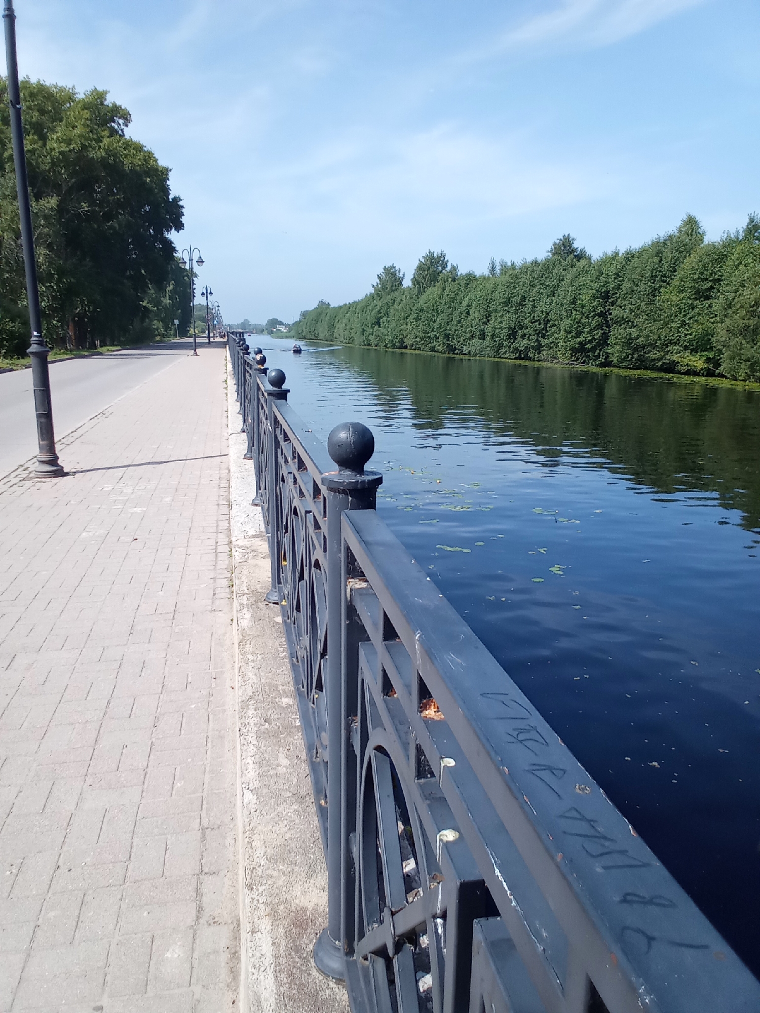 The image depicts a canal-side walkway in Belozersk, Russia. On the left, a paved sidewalk runs parallel to a road, bordered by trees and lampposts. A dark metal fence with decorative elements separates the walkway from the canal. The canal itself is dark blue and reflects the sky and the dense green foliage lining its opposite bank. A few small boats are visible on the water in the distance. The sky is clear and blue, suggesting fair weather. The overall scene is one of a peaceful urban waterfront environment.