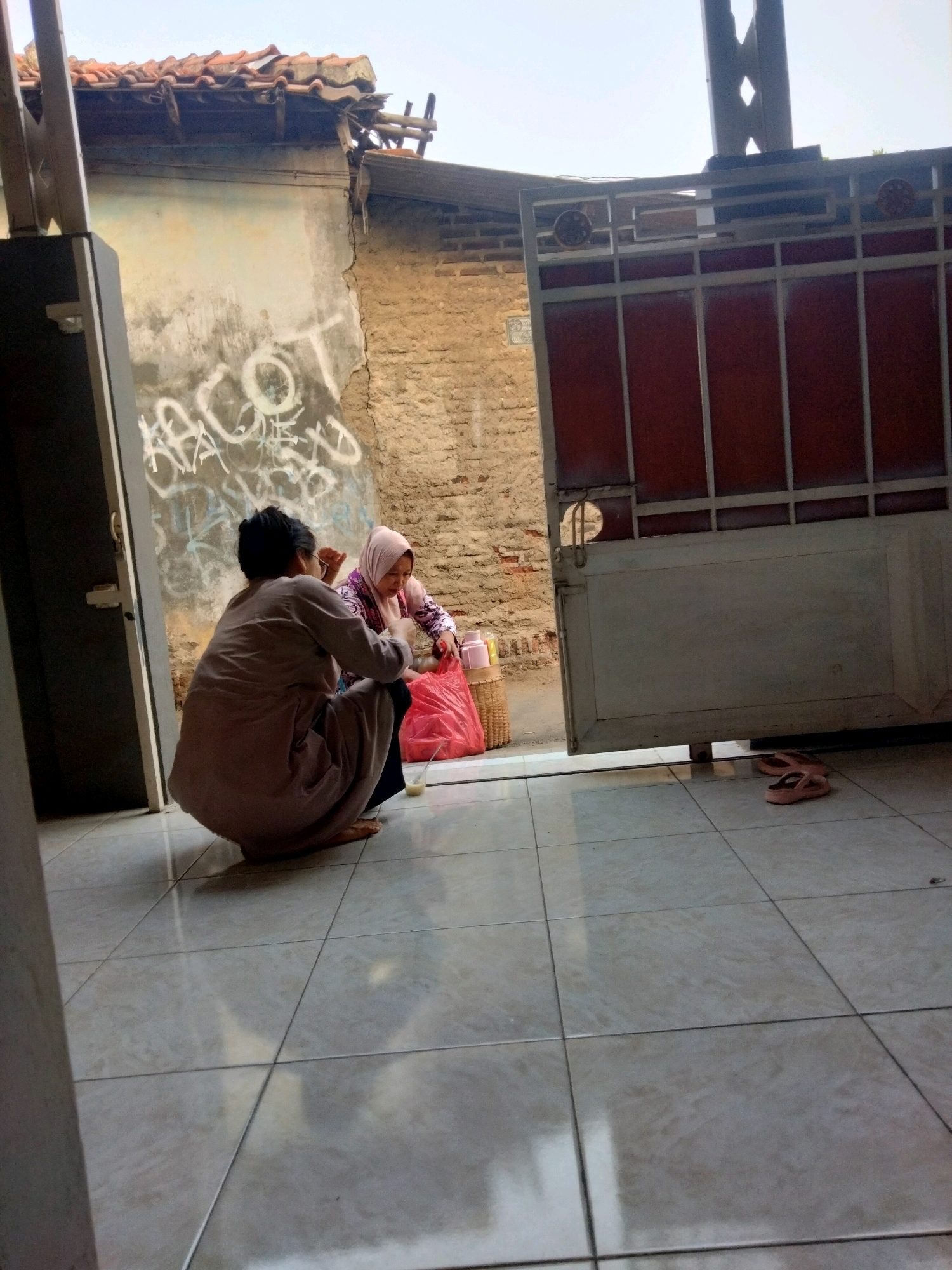 In the city of Suka Sari, Indonesia, two individuals are interacting on a tiled floor in front of an open gate. A person wearing a light brown robe is squatting, facing away from the viewer, while another person wearing a light pink hijab and a patterned top is also crouched down. In front of them, a large red plastic bag, a woven basket, and a pink thermos are visible. The background shows a weathered wall with graffiti and a reddish-brown brick wall, partially obscured by a dark metal gate. The environment suggests an outdoor or semi-outdoor setting, possibly a courtyard or entryway. The image captures a moment of interaction between the two people, with the objects suggesting they may be engaged in a transaction or sharing of items.