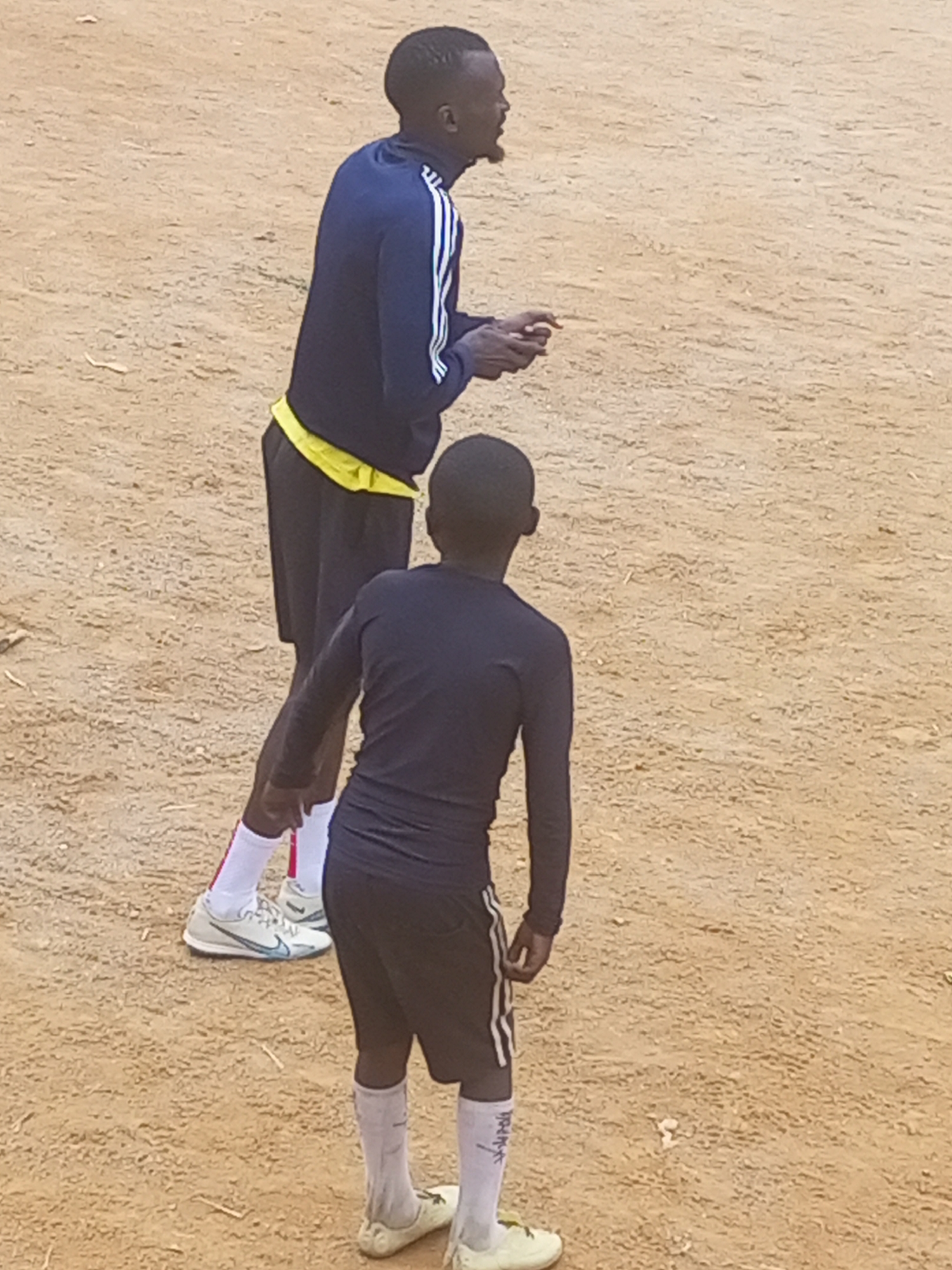 Two young Black males, likely teenagers, are standing on a dirt field. The male in the foreground is wearing a dark long-sleeved shirt with white vertical stripes on the sleeves, dark shorts with white stripes on the sides, and white socks with dark trim. The male in the background is wearing a dark long-sleeved shirt with white stripes on the shoulders and sleeves, black shorts with yellow trim, and white socks with red stripes. Both males appear to be engaged in a sporting activity, possibly soccer. The environment is a dry, dusty, outdoor area. The image is taken from a high angle, looking down at the subjects.