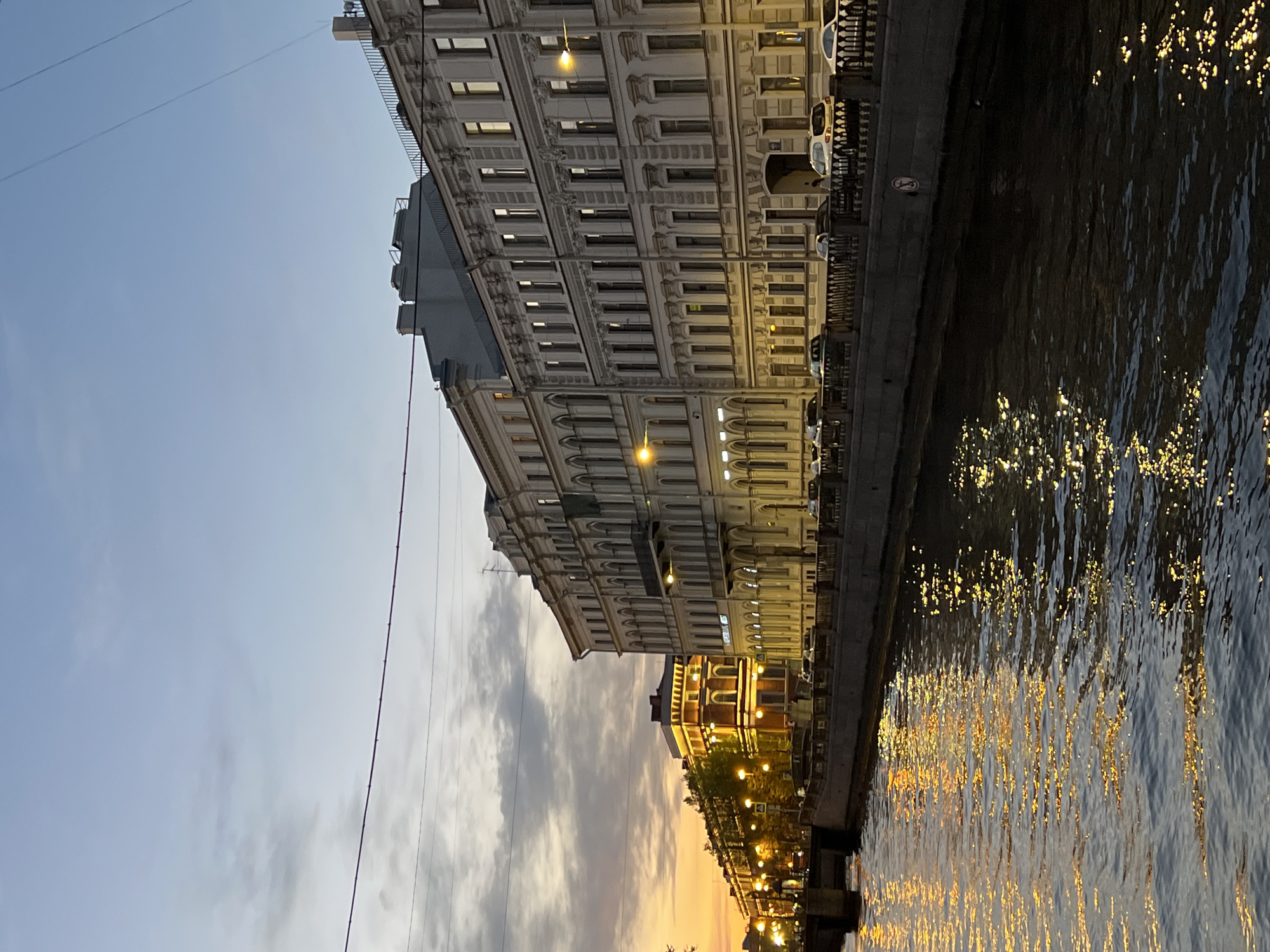 The image depicts a canal in Saint Petersburg, Russia, at dusk. On the left, an ornate building with multiple stories faces the canal, with streetlights illuminating its facade. Parked cars line the embankment along the canal's edge. The sky is a mix of pale blue and shades of orange and gray from the setting sun. The water reflects the city lights as golden ripples. Power lines stretch across the sky on the left side of the frame. Further down the canal, smaller buildings are visible.
