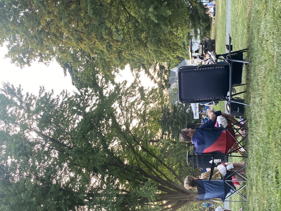 This image depicts an outdoor gathering in a grassy park area in Ridgefield, United States, during late afternoon or early evening. The scene is illuminated by sunlight filtering through the dense foliage of large, mature deciduous trees that frame the view.
In the foreground, three individuals are seated in portable chairs with their backs to the camera, facing an uncaptured central event or stage. From left to right:
1. An individual with light-colored hair is seated in a blue and black folding chair. A white bottle is visible on the ground next to the chair.
2. A male, wearing a blue polo shirt and light-colored shorts, is seated in a red, blue, and black folding chair. A dark bag or cooler rests on the grass near his right foot.
3. A large, black zero-gravity recliner chair is positioned, largely obscuring the person seated within it. A light-colored object, possibly a shoe, is visible on its footrest.
A black, slatted metal trash receptacle is positioned directly behind the two left-most seated individuals.
In the midground, a dark utility vehicle or golf cart, occupied by at least two individuals, is parked on the grassy field towards the right.
The background shows additional people scattered across the grass, some seated, suggesting an audience for an event. A white tent structure with dark lettering, possibly "CHIN," is visible on the left side of the background, indicating an organized gathering. Distant structures are faintly discernible beyond the tree line. The ground is covered in green grass, and the sky above is bright.