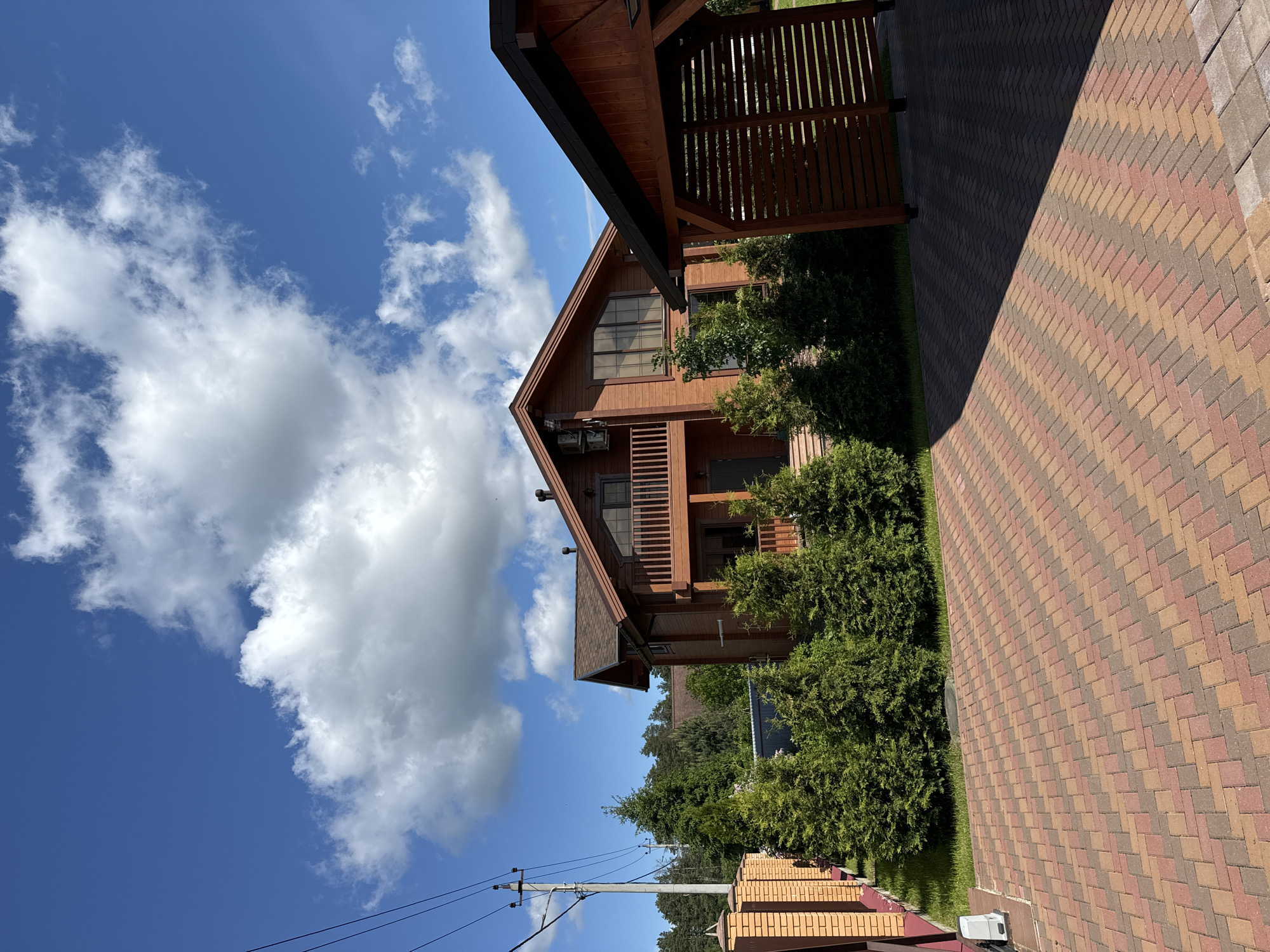 The image shows a residential scene in Vysokinichi, Russia. A large, wooden two-story house is the central focus, surrounded by manicured green bushes. A brick-paved walkway extends alongside the house. The sky is blue with scattered white clouds. Utility poles with power lines are visible in the lower left corner. A covered wooden structure is attached to the house on the right side. No people are visible in the image.