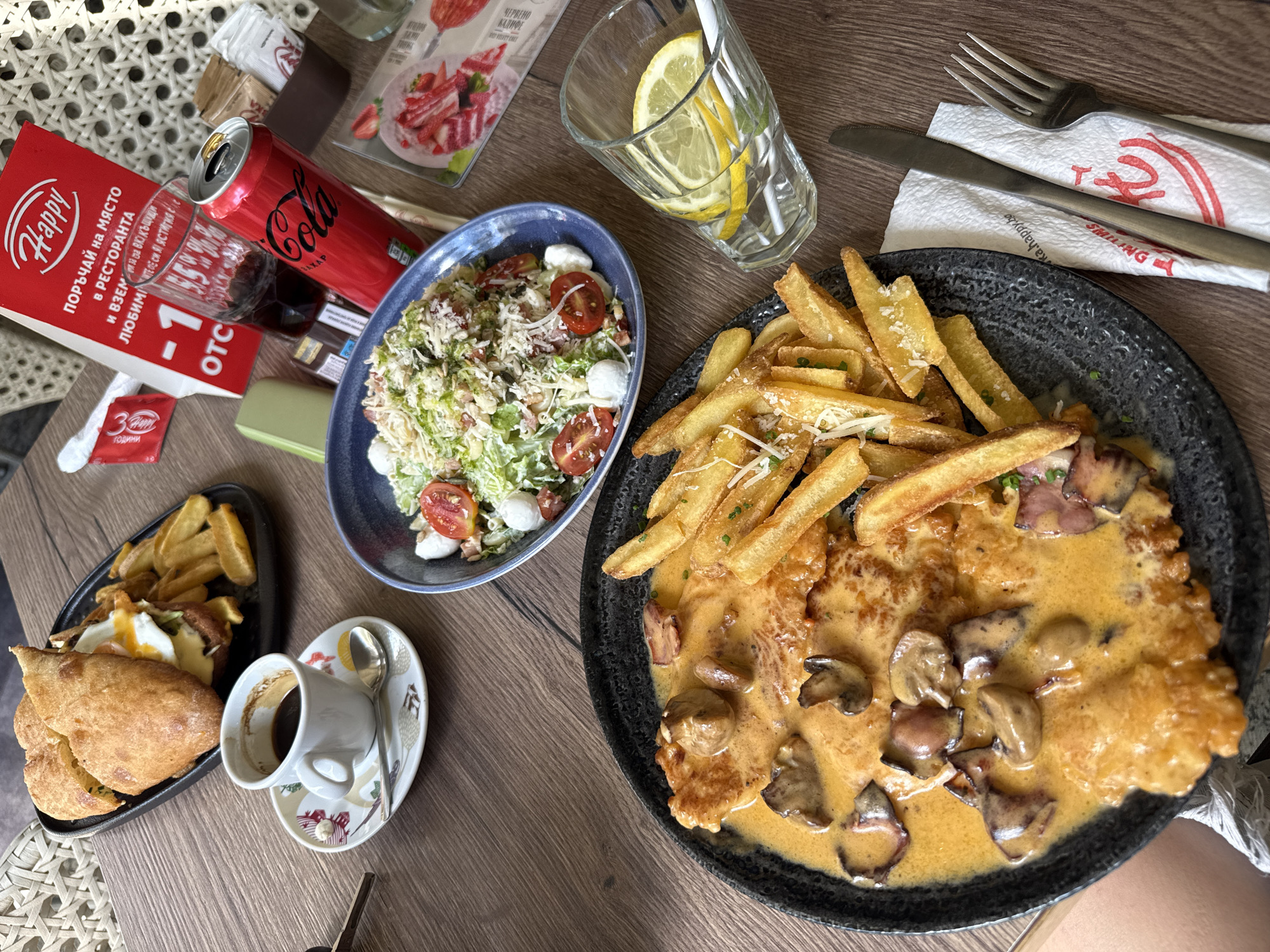 The image depicts a table setting at a restaurant. Dishes are scattered across the table, including a salad, fries, schnitzel with a mushroom sauce, and a plate of fried bread with fried eggs. Beverages consist of a Coca-Cola, a glass of water with lemon slices, and a cup of coffee. Silverware and napkins are also present. A "Happy" promotional item suggests that this is a Happy Bar & Grill restaurant. Given the Cyrillic script on the promotional material and the name of the city (Plovdiv) in Bulgaria, the scene is taking place at a Happy Bar & Grill location in Plovdiv, Bulgaria.