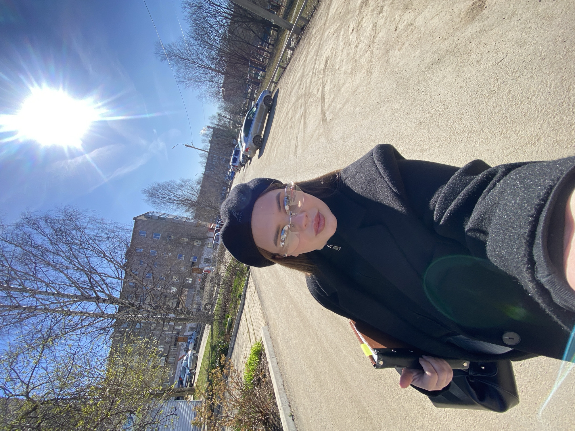A selfie taken on a sunny day in what appears to be Chernushka, Russia. The main subject is a person wearing a black beret, glasses, a black coat, and holding a black bag. They are standing on a paved street. In the background, there are apartment buildings, trees, and parked cars. The sun is bright in the upper left corner. The perspective is tilted, giving a rotated view of the scene.